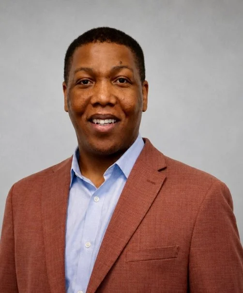 A man with short hair wearing a brown blazer and a light blue shirt, smiling against a plain light grey background.