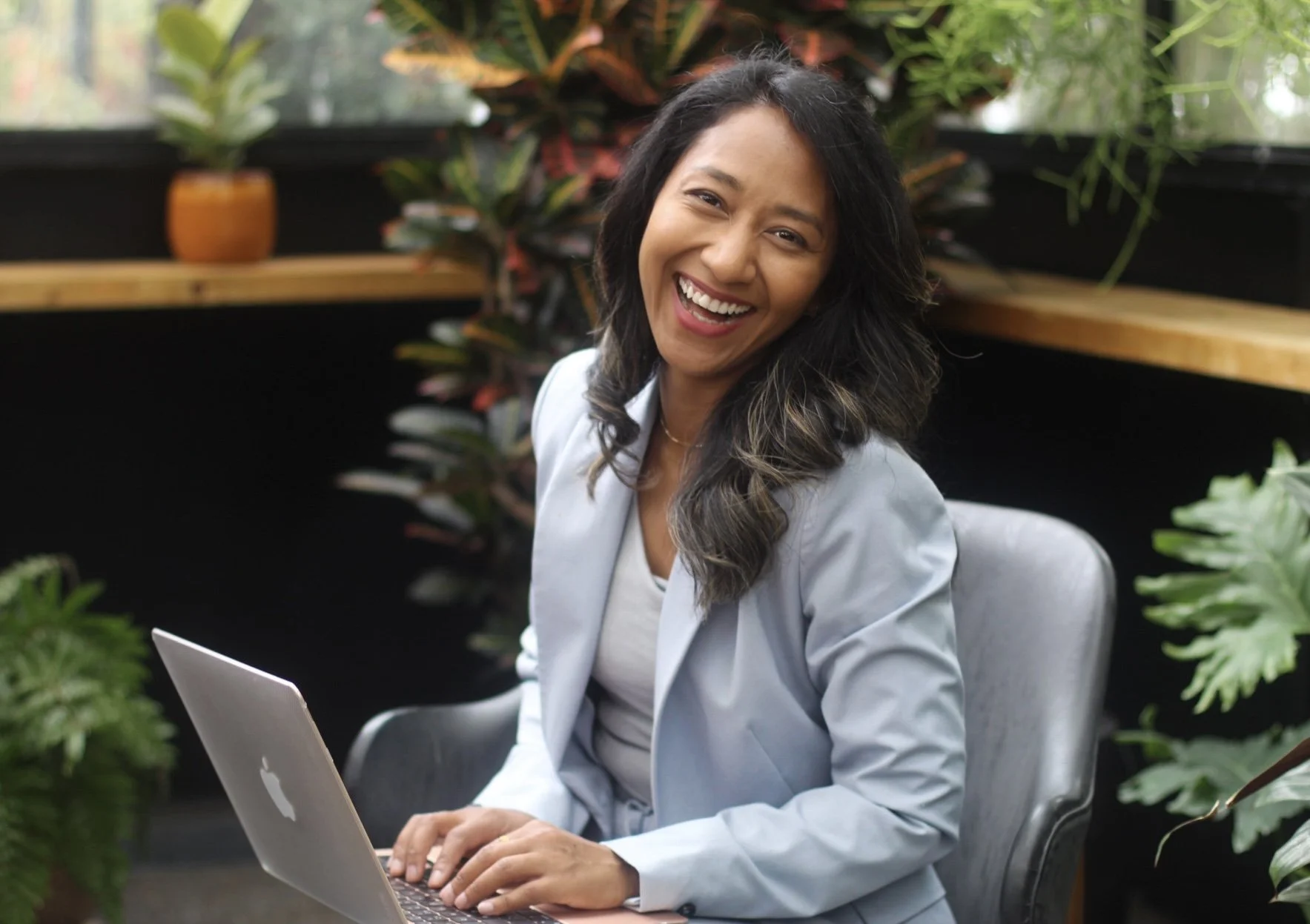 A woman with long dark hair smiling while working on a silver laptop in a bright indoor space surrounded by green plants.