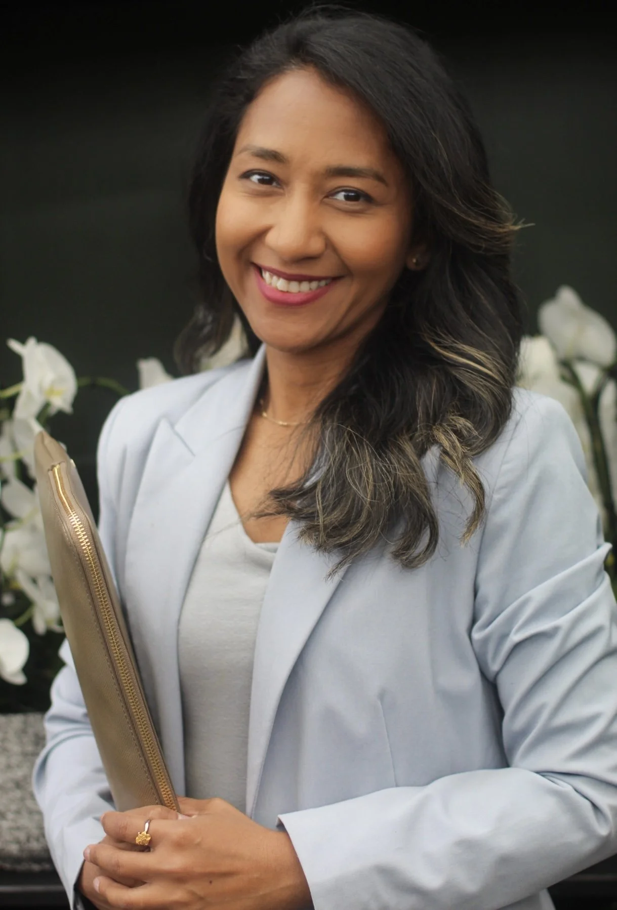 Portrait of a smiling woman with dark hair, wearing a light gray blazer, holding a beige folder, standing outdoors with white flowers in the background.