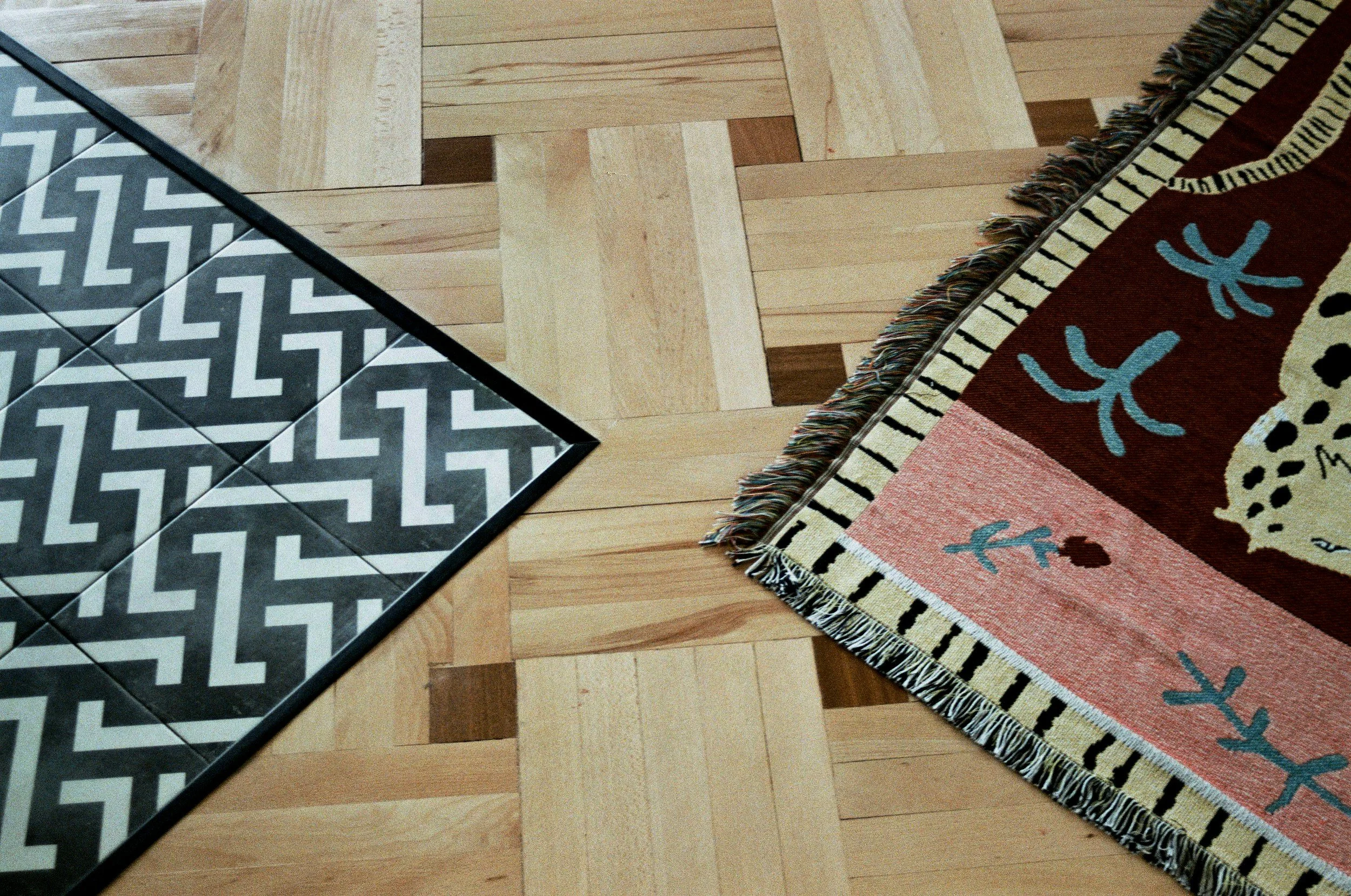 Part of a black and white geometric patterned tile floor on the left, tiled square on the right, and part of a colorful patterned rug with fringes on a wooden floor.