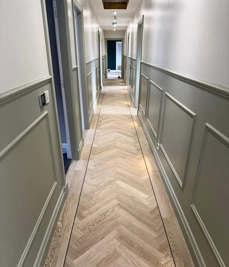 A long, narrow hallway with cream-colored walls, decorative wainscoting, and light wood herringbone patterned flooring, with a door at the end.