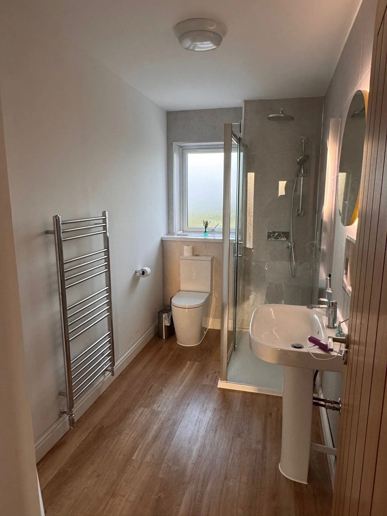 A bathroom with wooden flooring, a white toilet by a small window, a freestanding sink, a shower with a glass door, a wall-mounted showerhead, and a mirror. A heated towel rail is on the left wall, and a small green plant sits on the window sill.