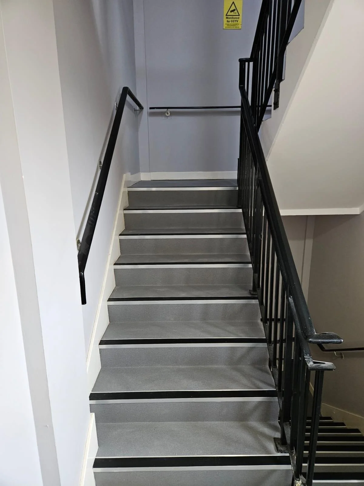 Indoor staircase with gray steps, black metal handrails on both sides, and a yellow warning sign on the wall at the top.