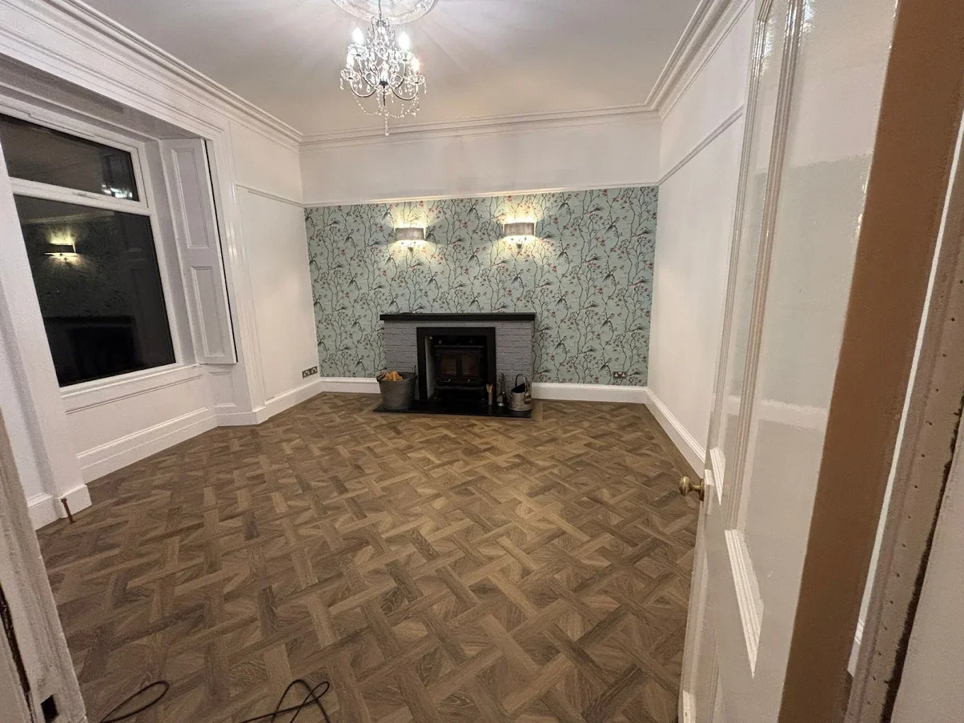 Empty living room with parquet wood floor, white walls, a fireplace with a grey brick surround, wallpapered accent wall, a chandelier hanging from the ceiling, and wall-mounted lights above the fireplace.