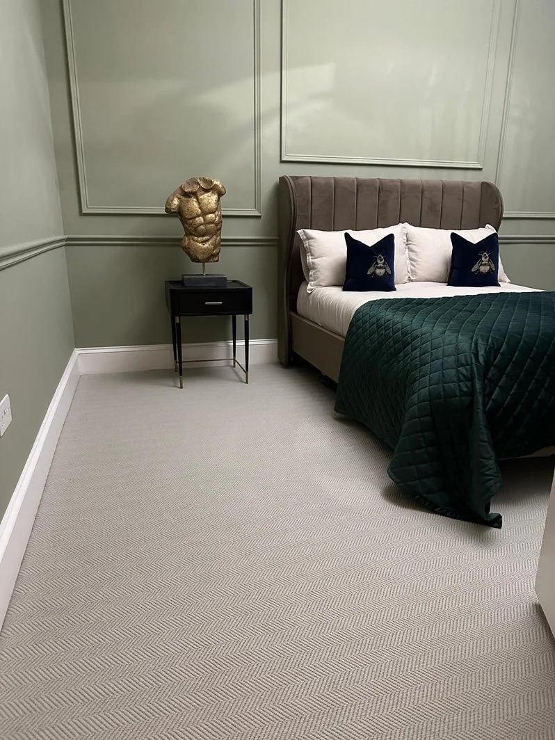 A bedroom with gray walls, a tall upholstered bed with white and navy pillows featuring bee embroidery, a dark green quilt, and a black side table with a decorative gold torso sculpture.