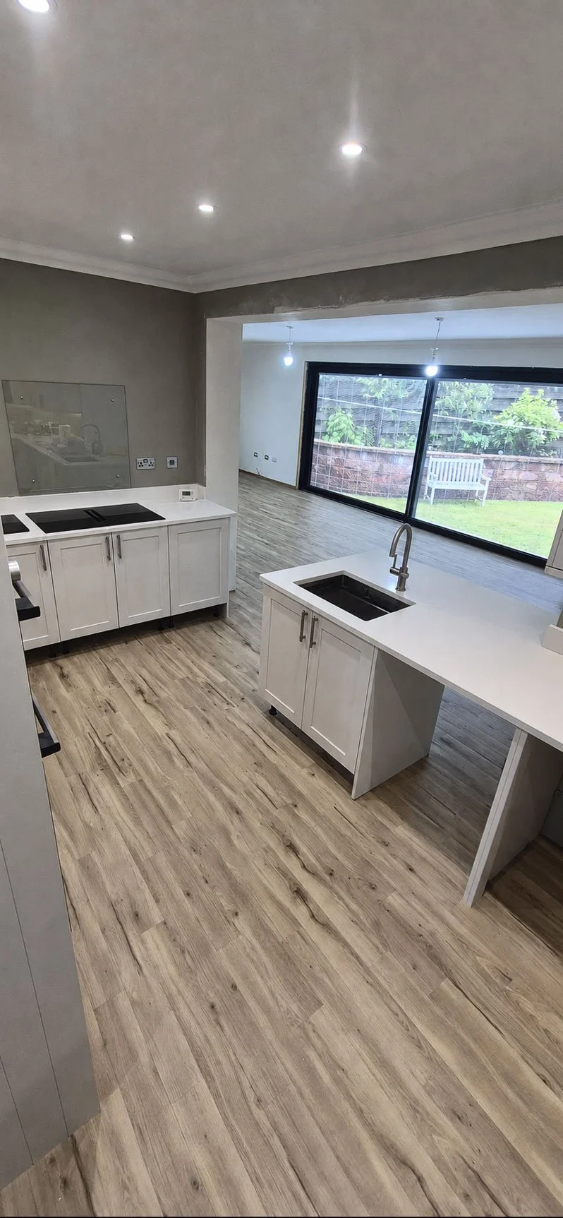 Modern kitchen with white cabinets, black cooktop, and a small island with a sink, open to a living area with large windows and a backyard garden.