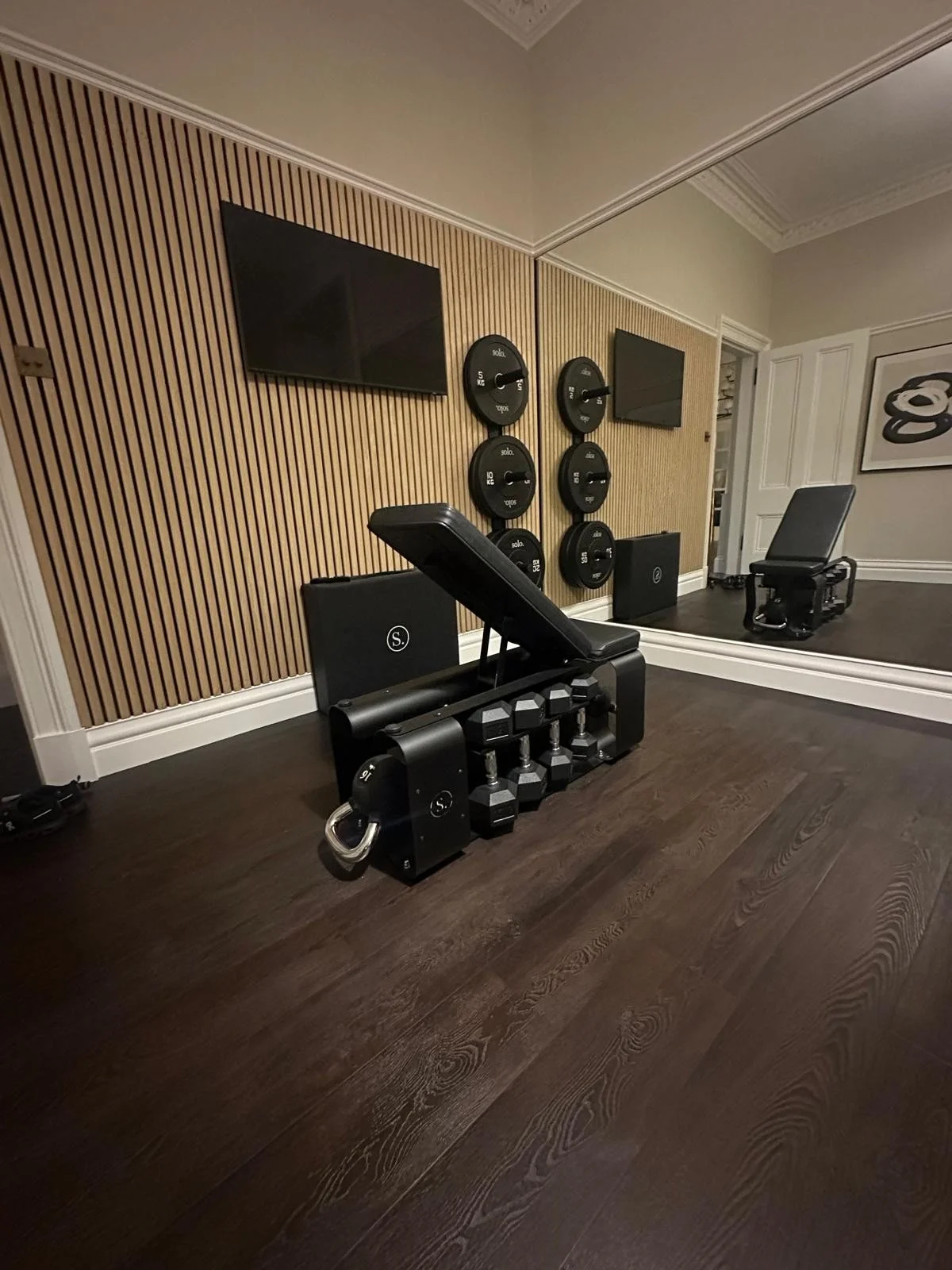 Home gym with two black workout chairs, dumbbells, wall-mounted weights, a mirror, and a wooden accent wall.