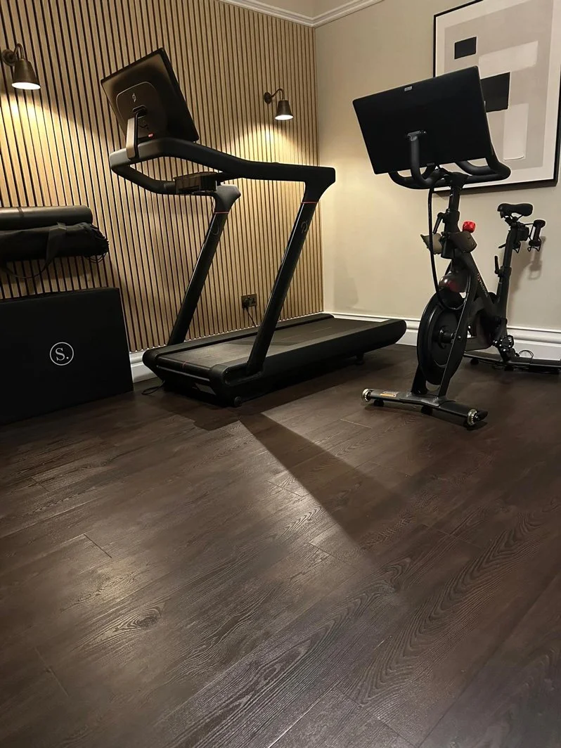A home gym with a treadmill and a stationary exercise bike on a dark wooden floor, with modern wall lighting and framed artwork.