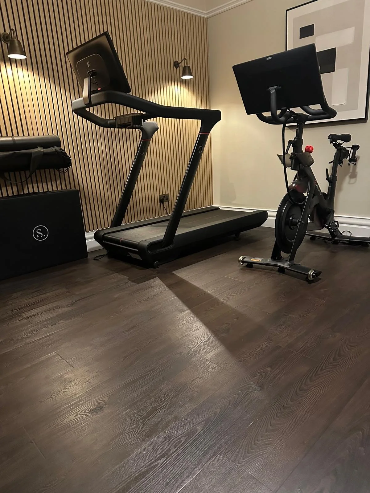 A workout room with a treadmill and an exercise bike, both positioned on a dark wood floor with a wooden slat wall and abstract artwork on a white wall.