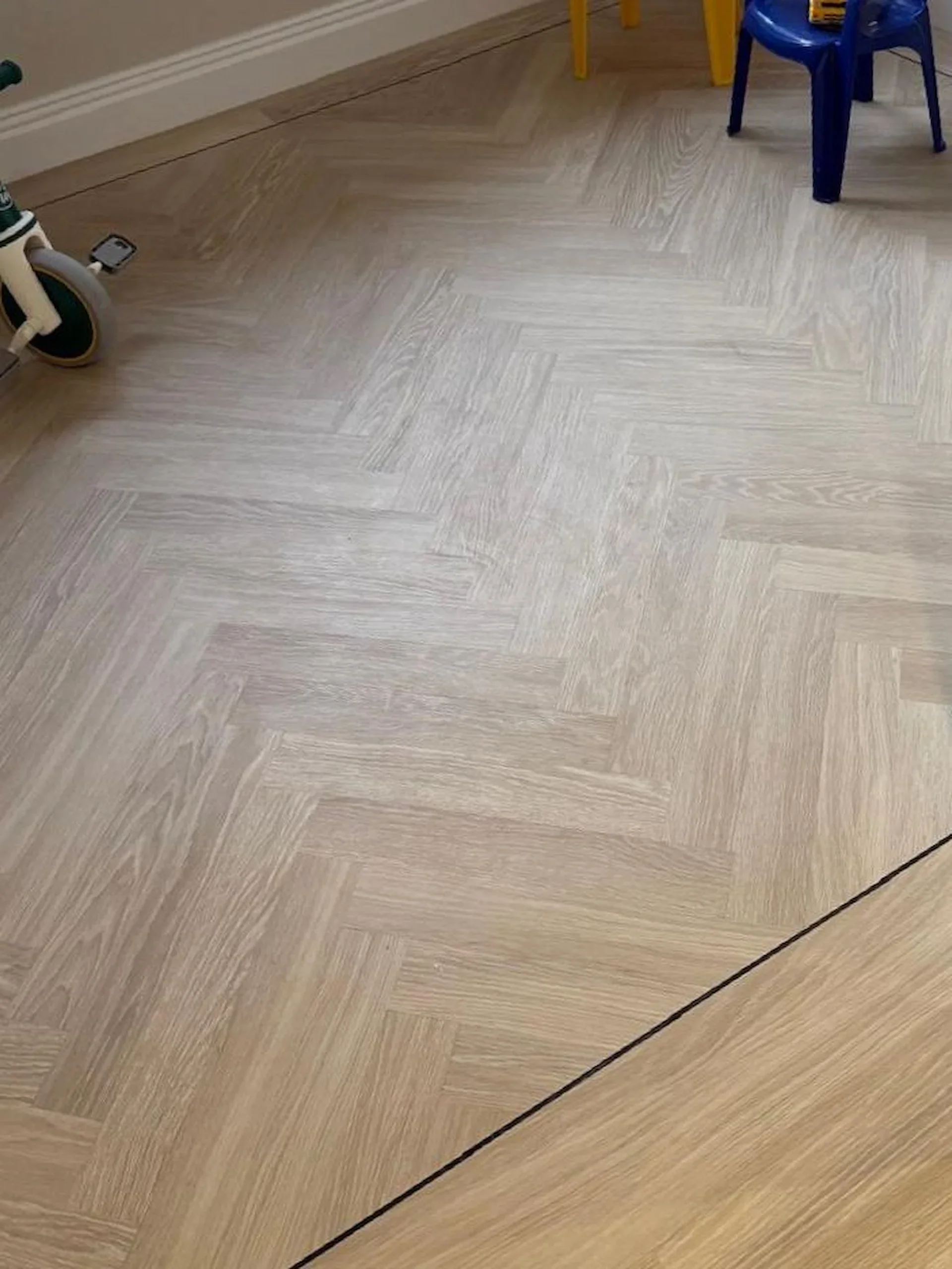Close-up of a herringbone-patterned wooden floor with a black line marking the boundary and a portion of a child's blue chair, yellow furniture, and a toy scooter visible.