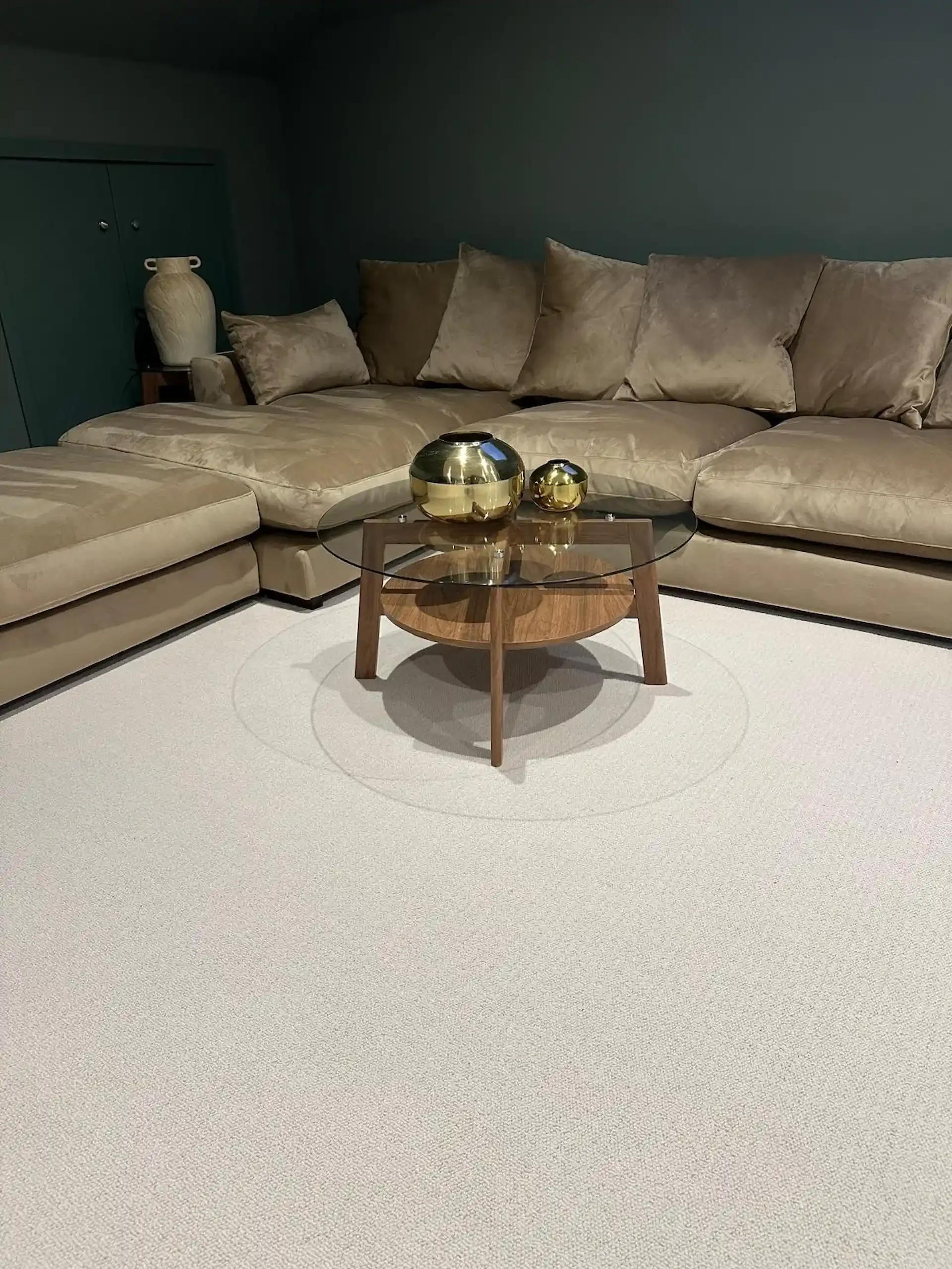 Living room with beige sectional sofa, round wooden coffee table with gold vases, dark green walls, and a large beige decorative vase on a side table.