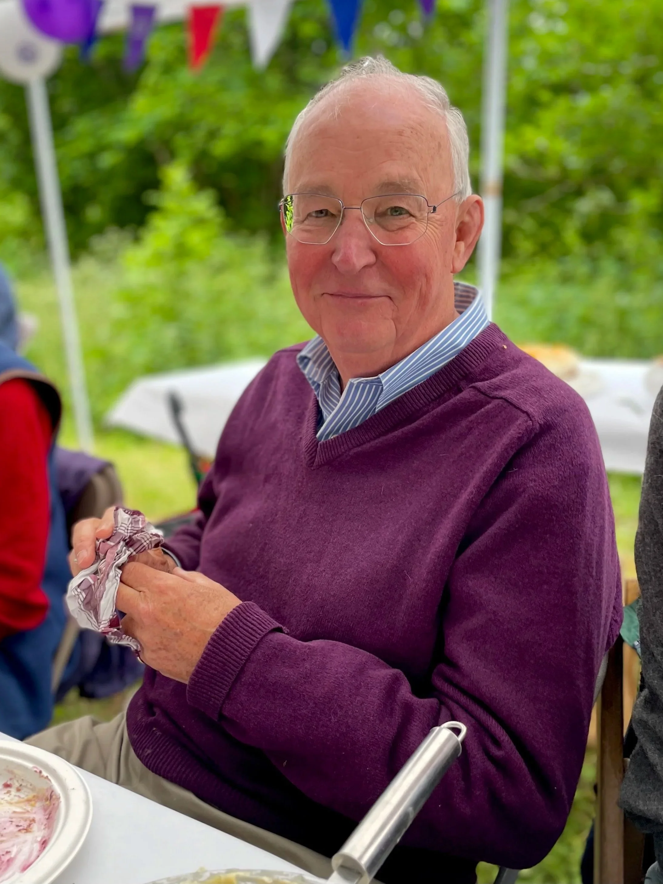 An elderly man with glasses and gray hair smiling at a gathering outdoors, wearing a purple sweater over a blue striped shirt, with bunting decorations and green trees in the background.