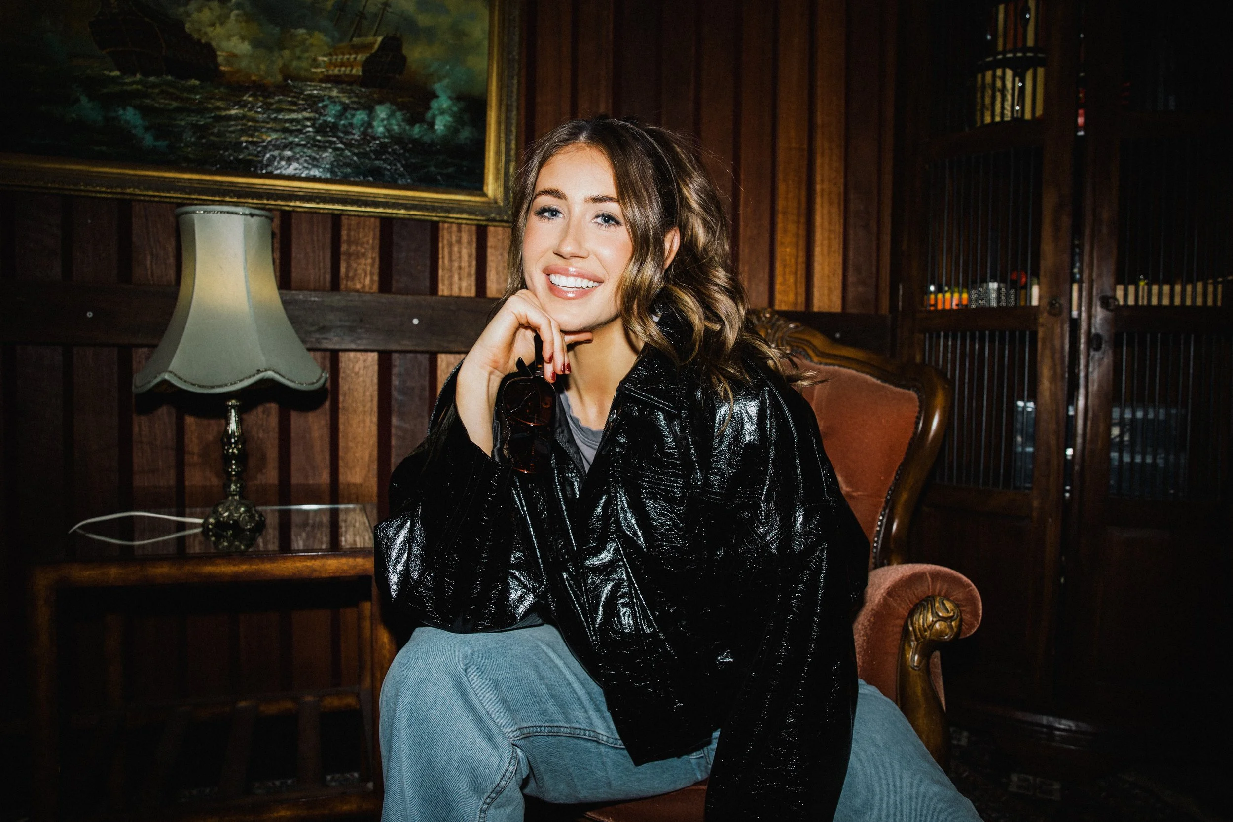 A young woman with brown wavy hair, wearing a shiny black jacket and light blue jeans, sitting on a wooden armchair with a pinkish upholstery. She has a bright smile, and her hand is resting on her chin. The background features a framed painting of a ship at sea, a vintage-style table lamp, wooden paneling, and a decorative wooden gate.