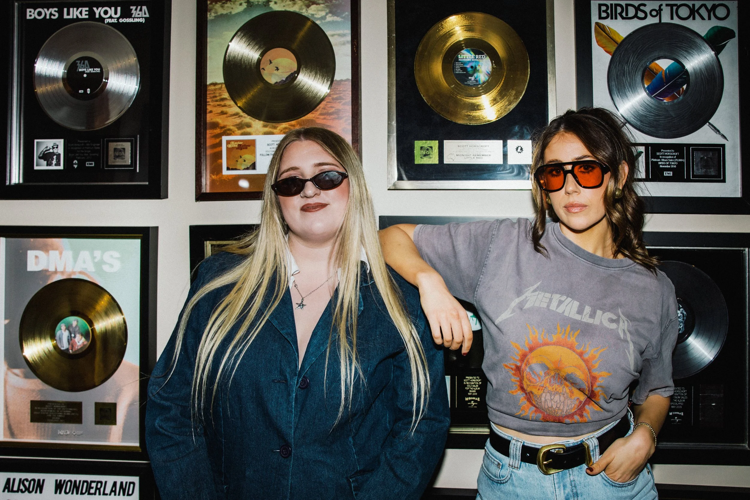 Two women wearing sunglasses pose in front of a wall decorated with framed gold records and album covers. One woman has long blonde hair and the other has shoulder-length dark hair.