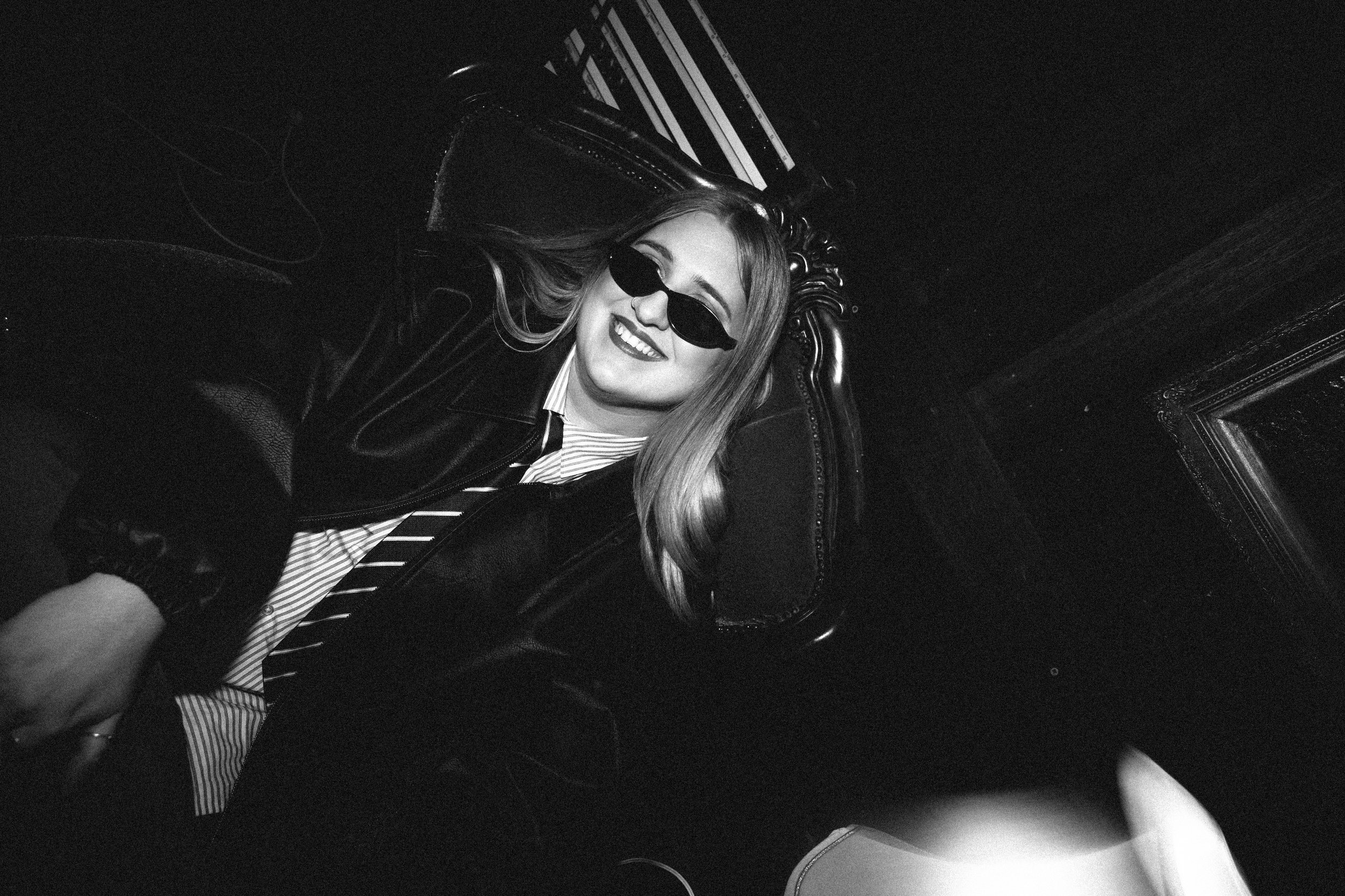 A black-and-white photo of a smiling woman with long hair, wearing sunglasses and a striped shirt, sitting in a vintage leather chair.