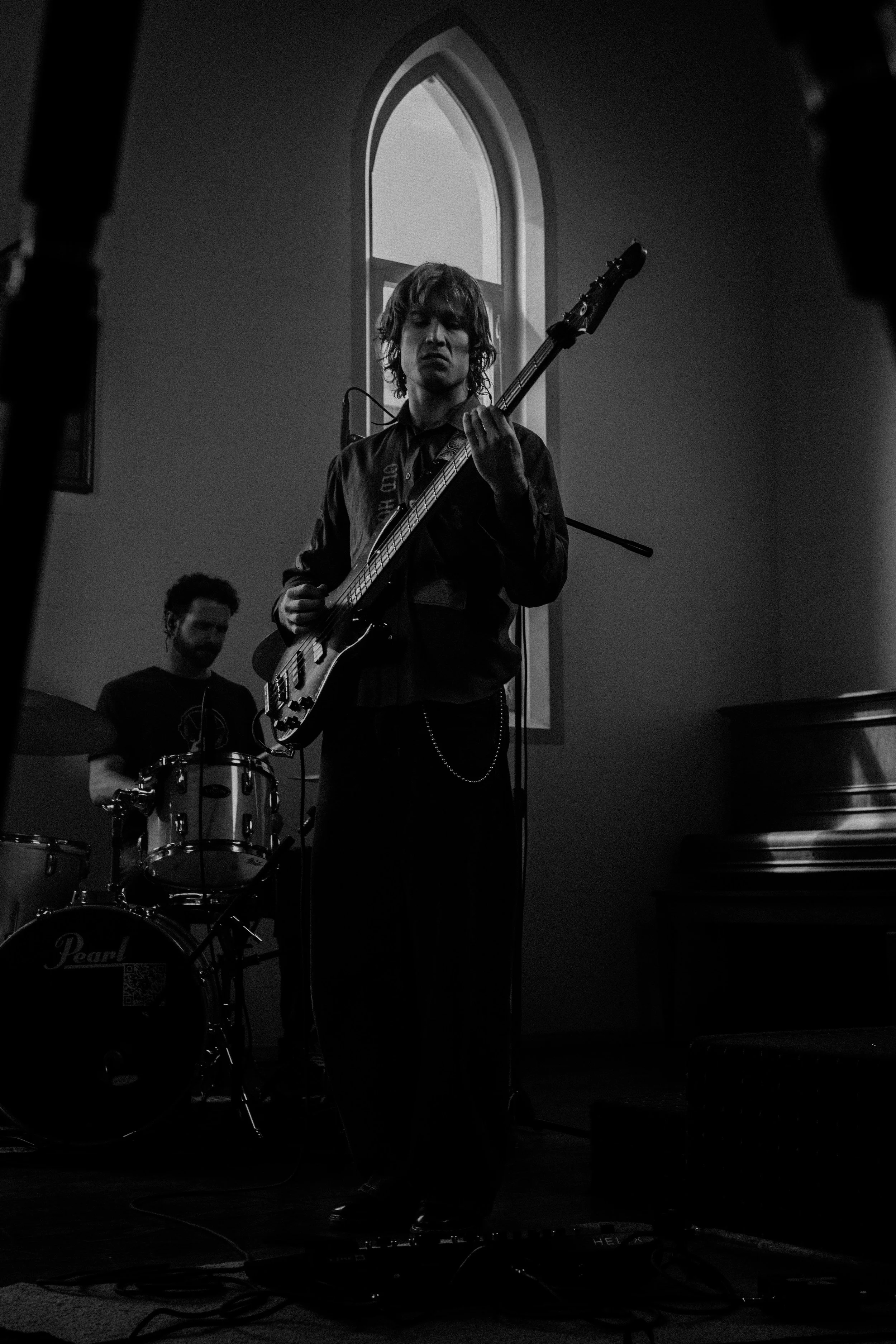 Black and white photo of a young man playing an electric guitar and singing into a microphone, with a drummer in the background inside a church with a tall arched window.