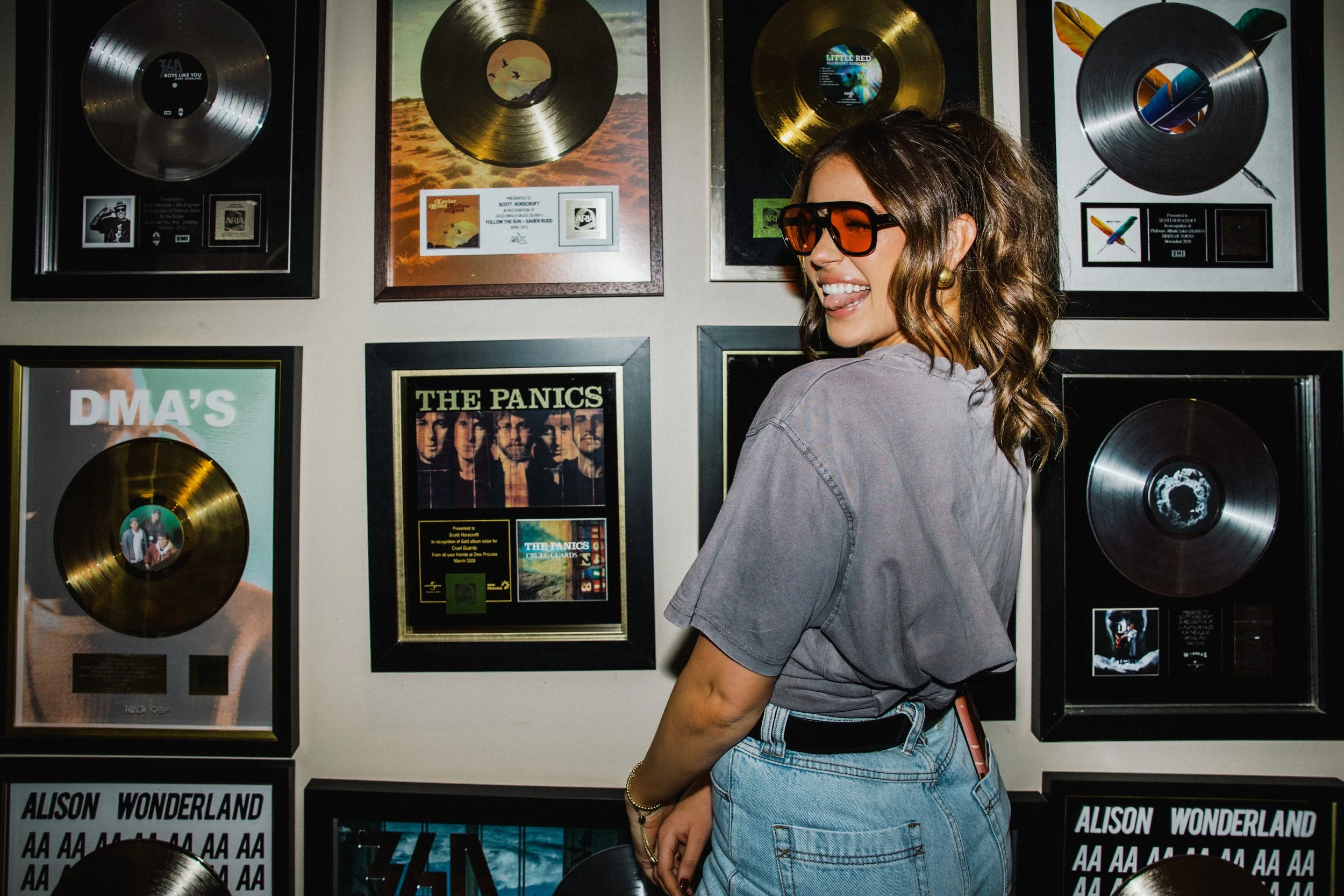 A woman with shoulder-length brown hair wearing large orange sunglasses, a gray T-shirt, and light blue jeans, is smiling and sticking her tongue out while looking to her right. She is standing in front of a wall decorated with various gold record awards and framed album covers, including one titled "The Panic" and another titled "D.M.A's."