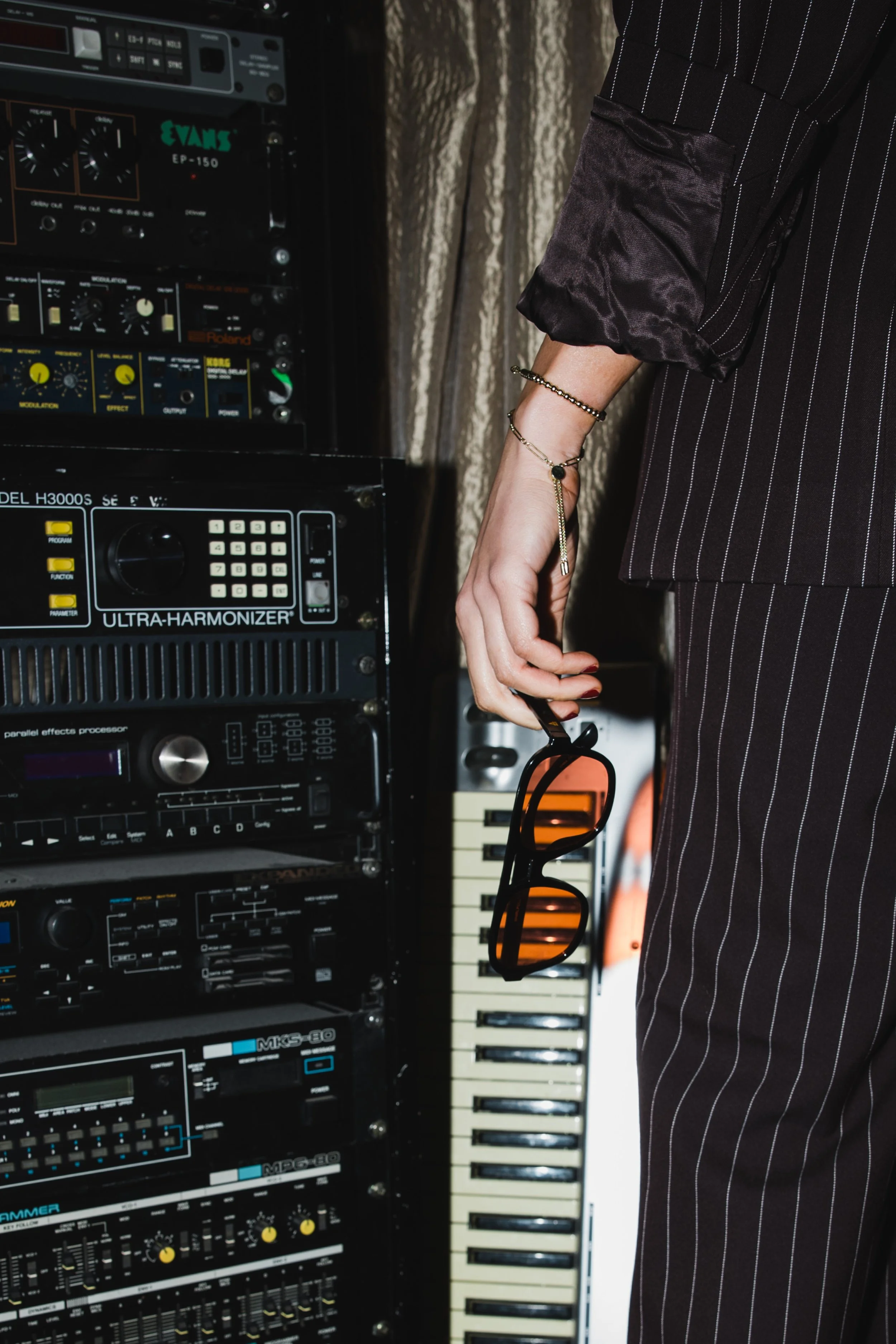 Person wearing a black pinstripe suit holding sunglasses, standing near audio equipment and a keyboard.