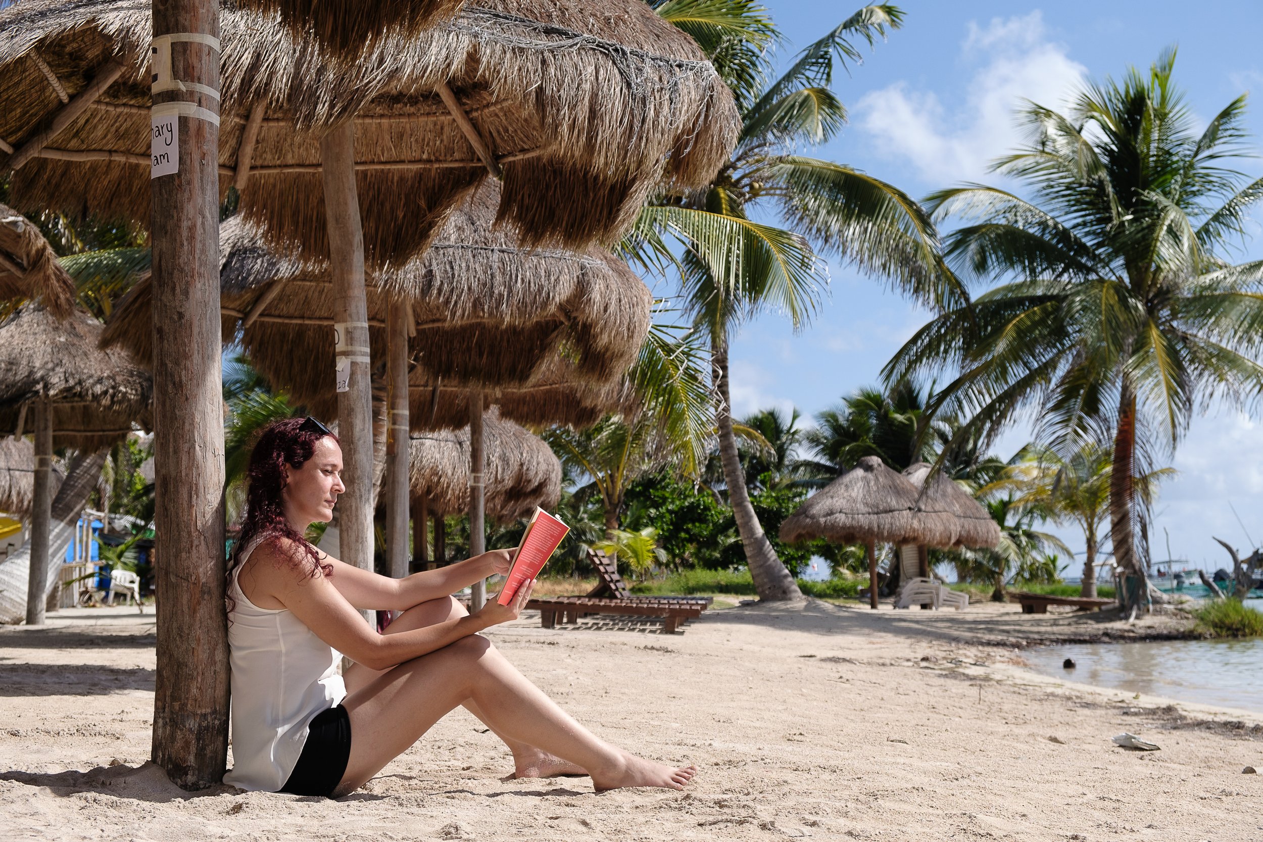 A woman sitting on a sandy beach under a thatched umbrella, reading a book with palm trees and blue sky in the background.