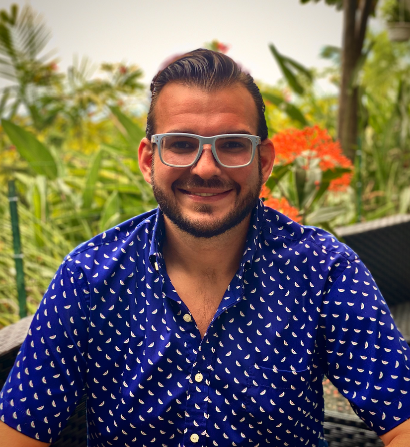 A man with glasses smiling outdoors, with lush green plants and orange flowers in the background.