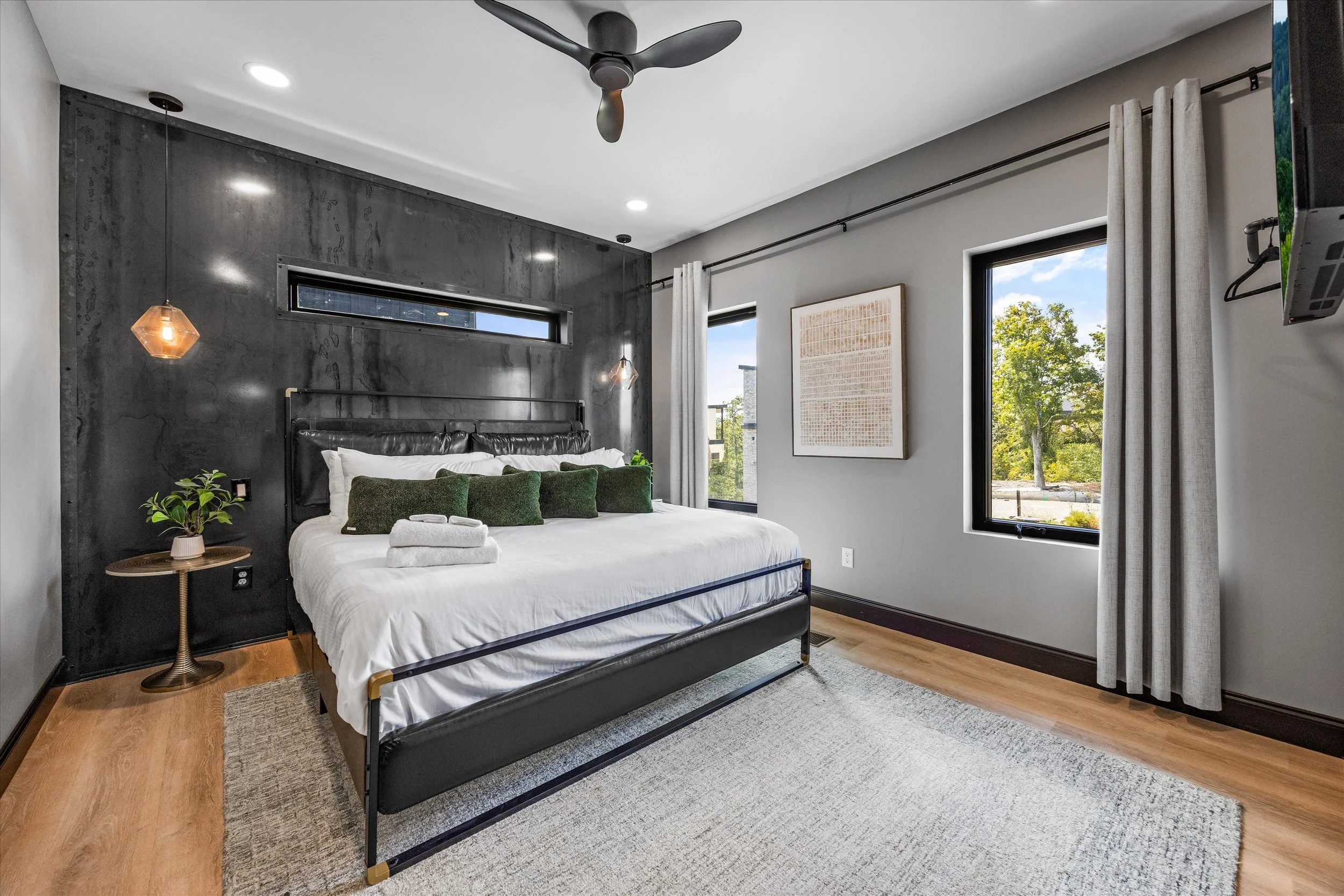 A modern bedroom featuring a black metal bed frame with white bedding and green pillows. There are two pendant lights hanging on either side of the bed, a small side table with a potted plant, and large windows with white curtains showing trees outside. The ceiling has a black ceiling fan and recessed lighting, with a textured dark accent wall behind the bed.
