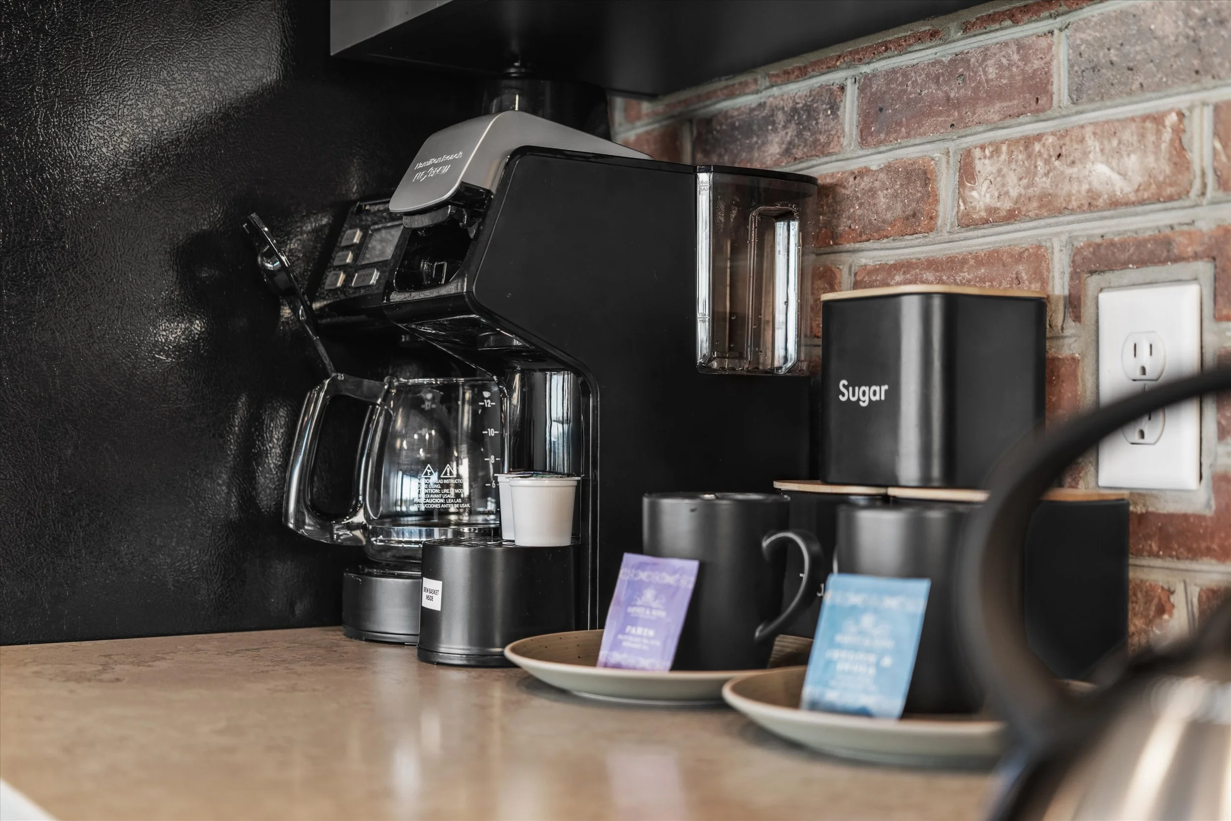 Black coffee maker on a kitchen counter with a glass carafe, next to black containers labeled 'Sugar,' and plates with information cards, against a brick wall background.