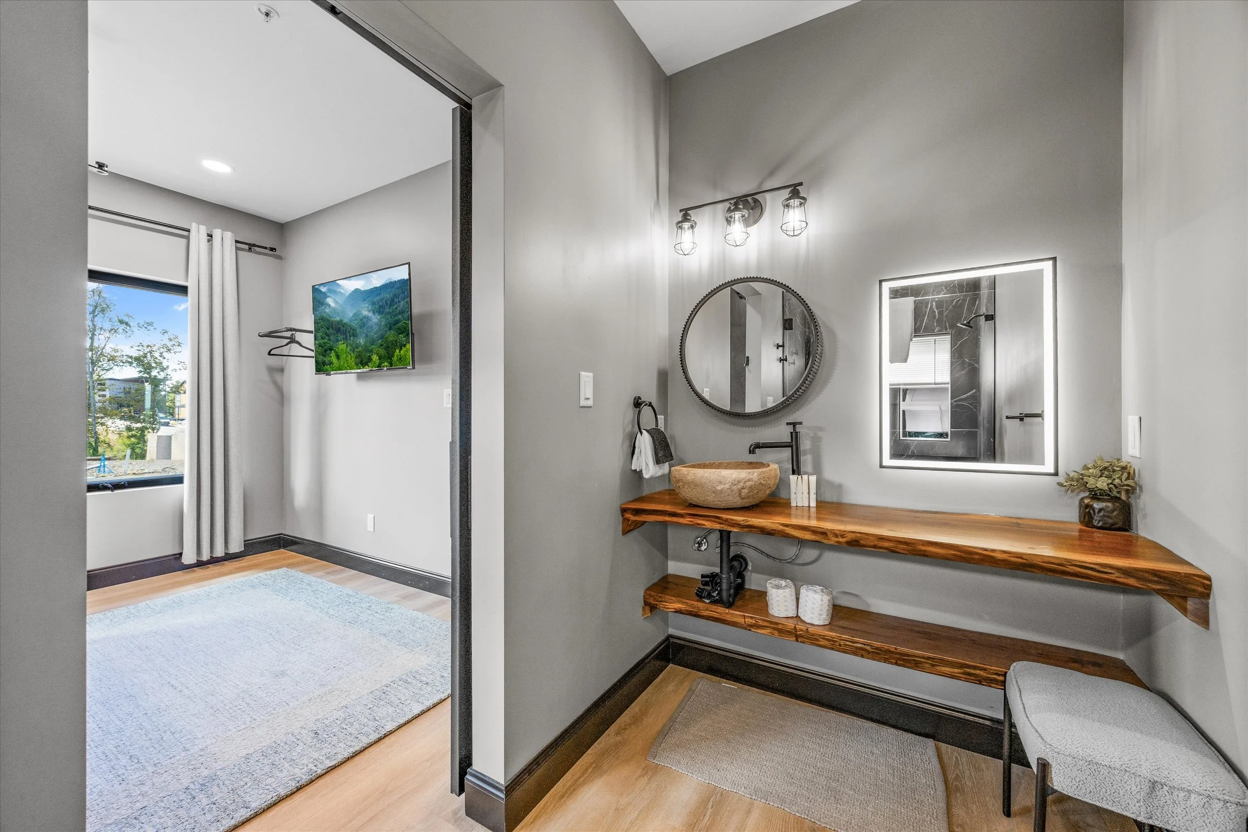 Modern bathroom with a floating wooden vanity, stone vessel sink, round mirror, wall-mounted light fixture, and a small potted plant. Adjacent laundry or dressing area with a window, TV, and hanging clothes.