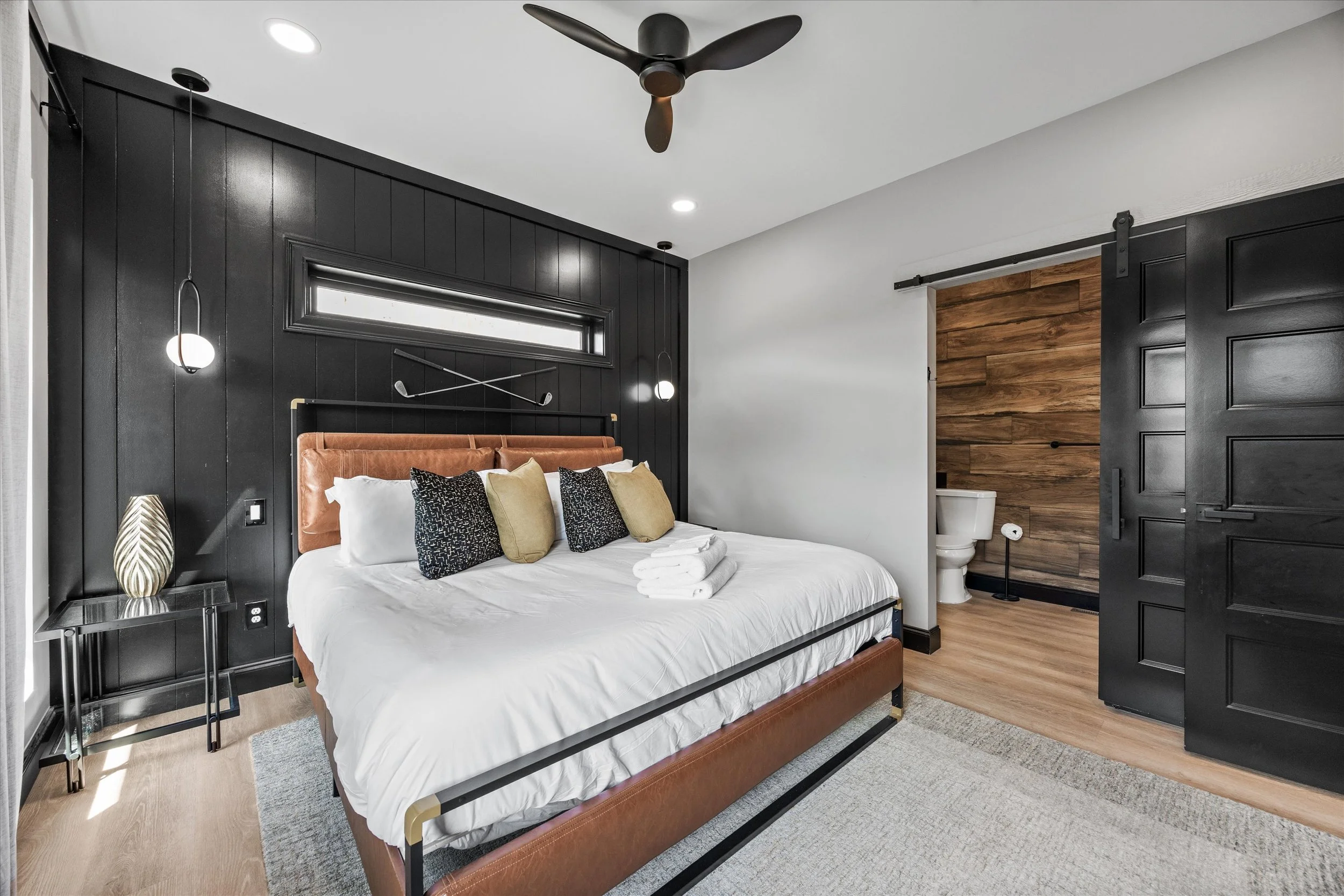Modern bedroom with black wood-paneled wall, brass bed frame, white bedding, decorative cushions, and a small bathroom with wooden wall accents visible through a sliding black barn door.