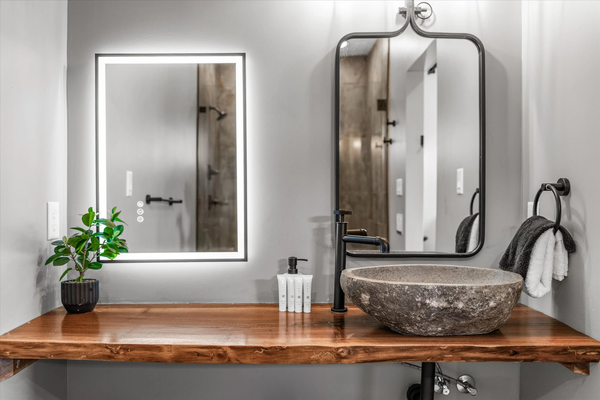 Modern bathroom with dual frameless mirrors, stone vessel sink, black fixtures, towel hooks with towels, green plant in black pot, white wall, and wooden countertop.