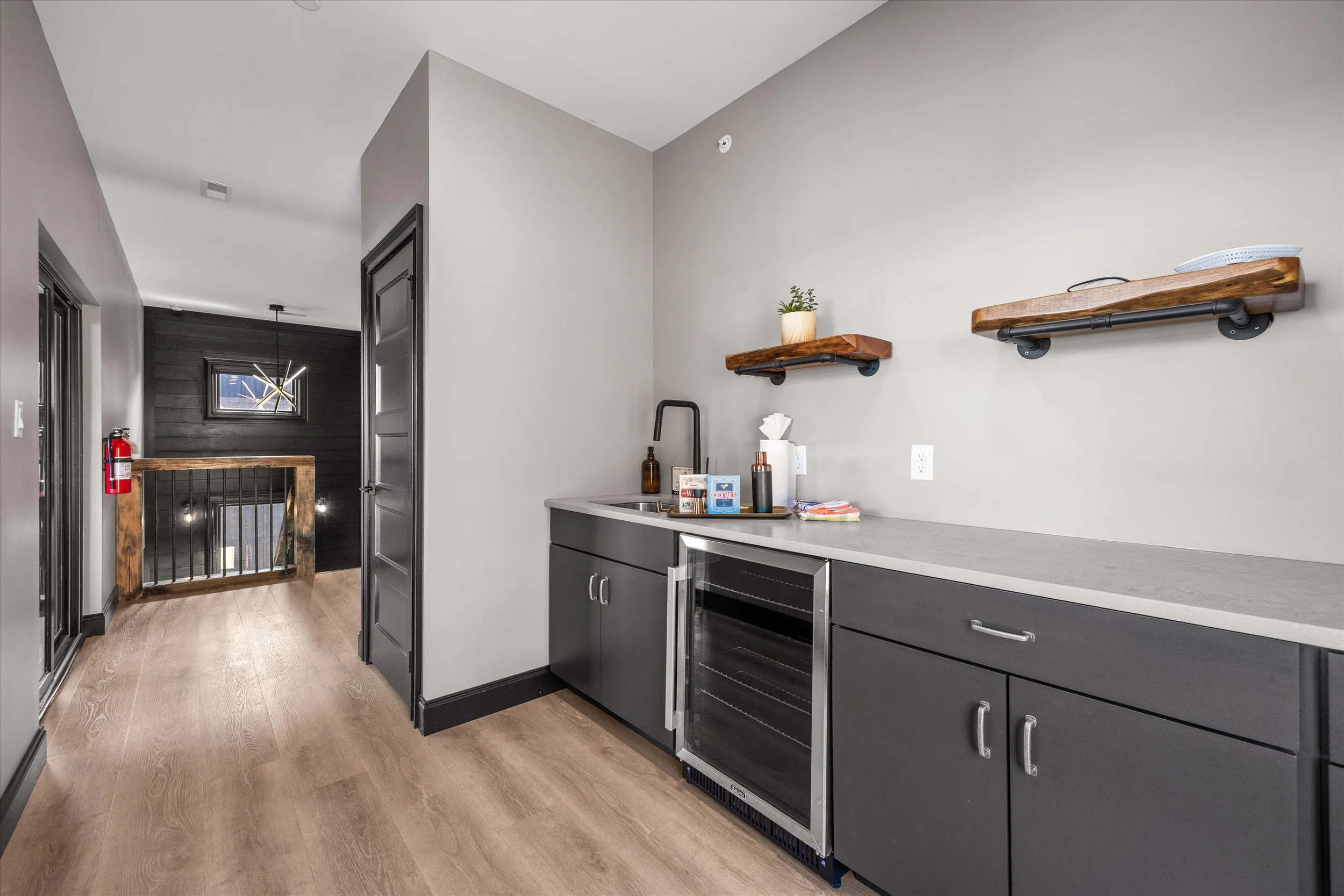 Modern kitchen with grey cabinets, a countertop with various items, and wall-mounted wooden shelves with piping detail. The kitchen is adjacent to a hallway with a wooden staircase and black walls.