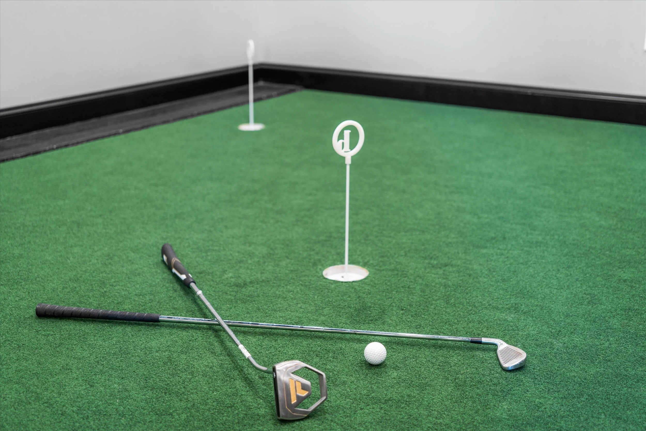 Miniature golf putting green with two golf clubs and a golf ball, small flags marking holes, and a black border on a gray wall.