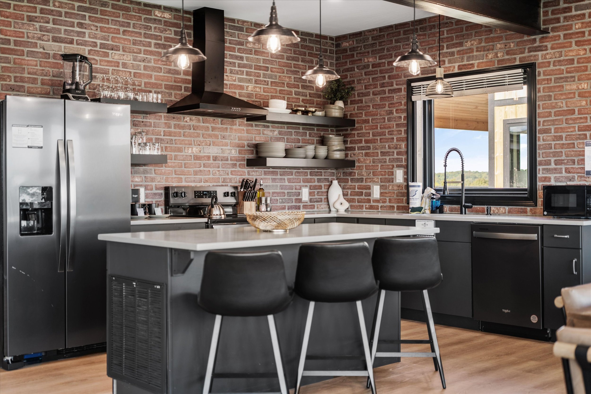 Modern kitchen with brick walls, black cabinets, stainless steel appliances, open shelving with dishes, and pendant lights over a white island.