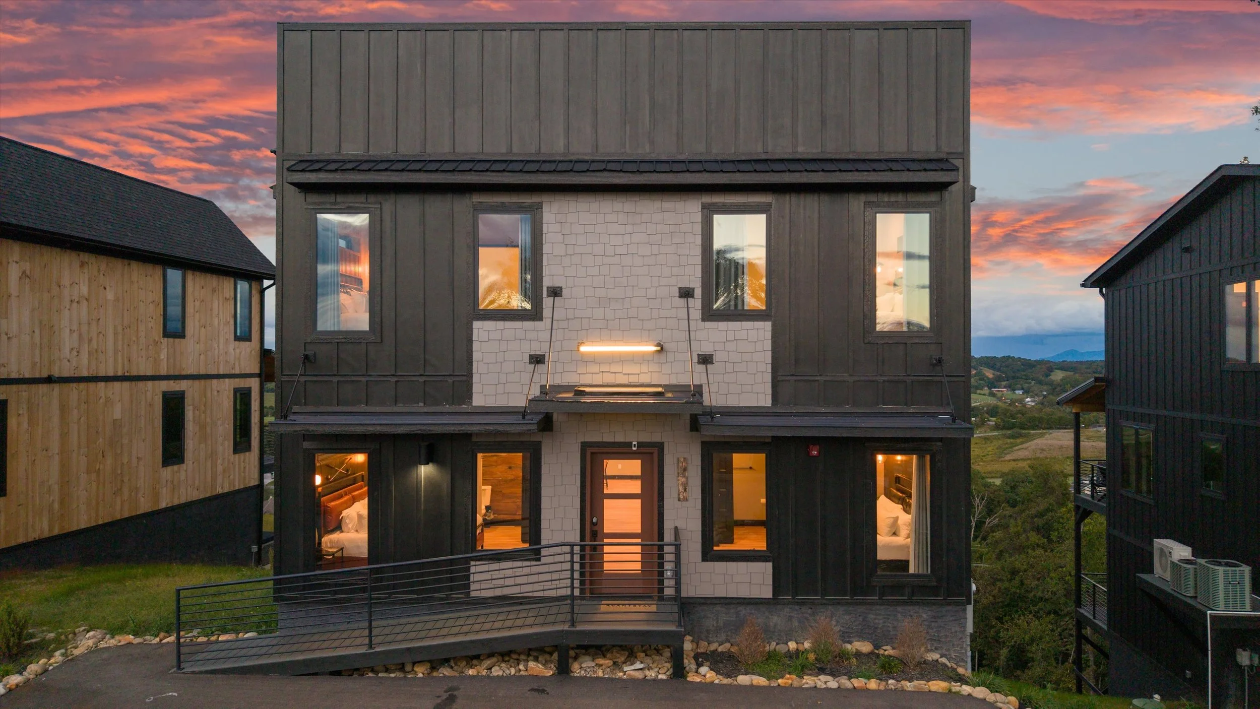 A modern multi-story house with black and white exterior, large windows reflecting the sunset sky, and a small balcony with a railing, surrounded by neighboring houses and natural landscape in the background.