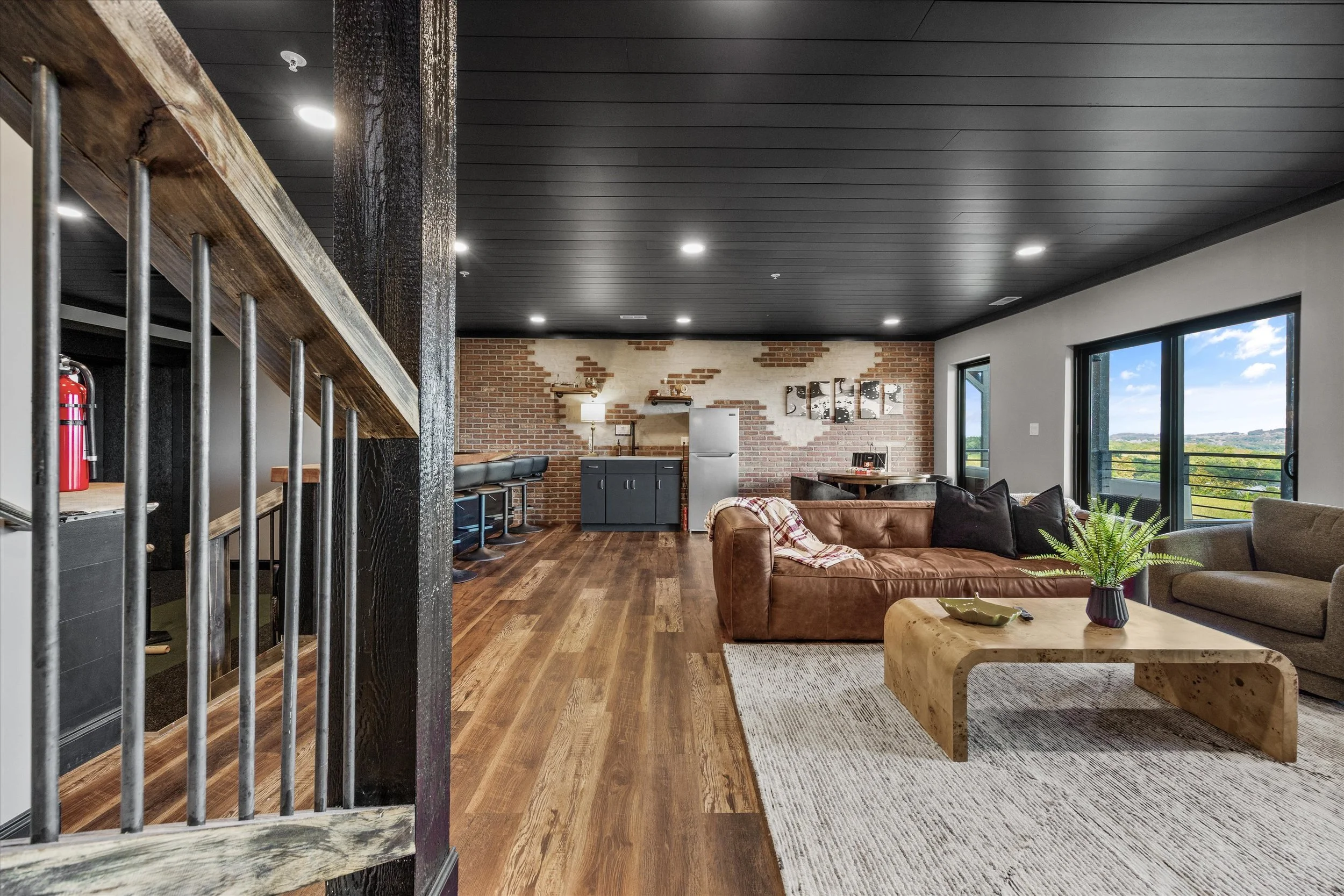 Open-plan living space with a brick wall, leather sofa, wooden coffee table, floor-to-ceiling windows, kitchen area with barstools, and a black ceiling with recessed lighting.