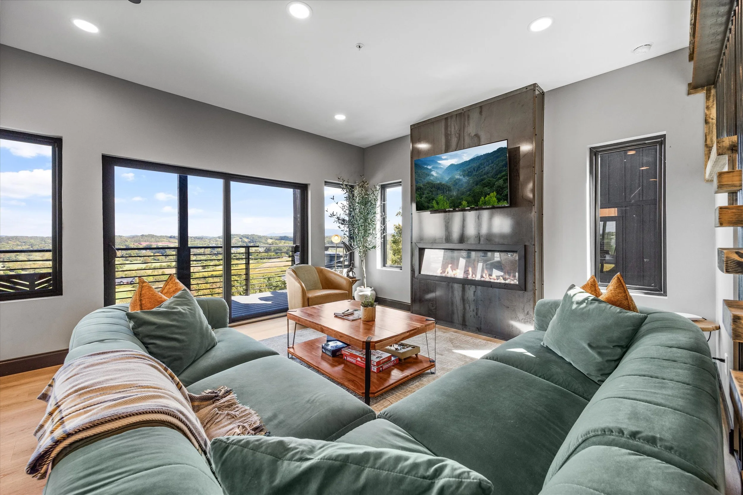 Living room with large window and balcony view of a mountain landscape, green sofa, armchair, wooden coffee table with books and game, fireplace, and mounted flat-screen TV.