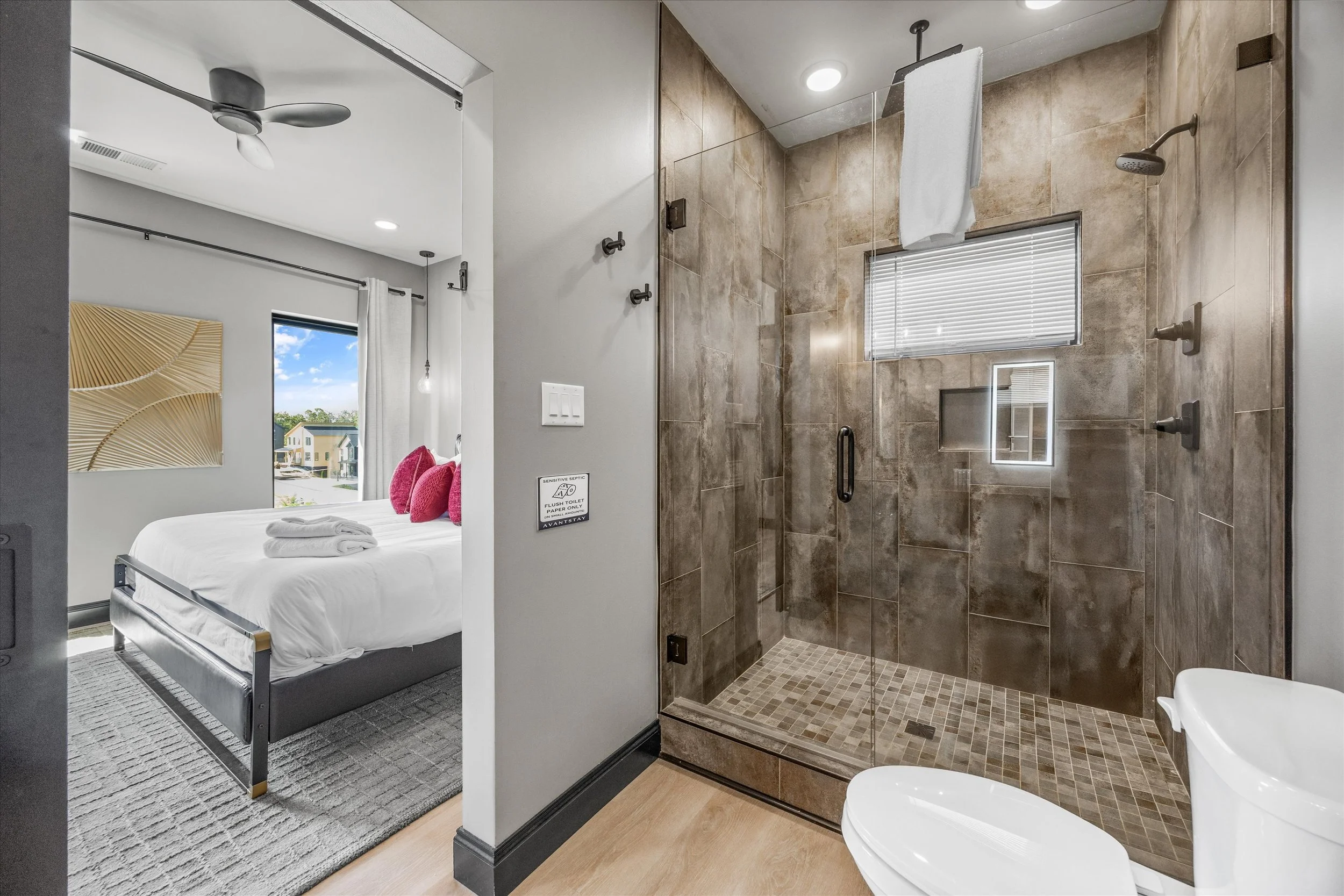 A room with a bed, window, and wall art on the left, and a walk-in shower with brown tiles, a small window, and a towel hanging from the showerhead on the right.