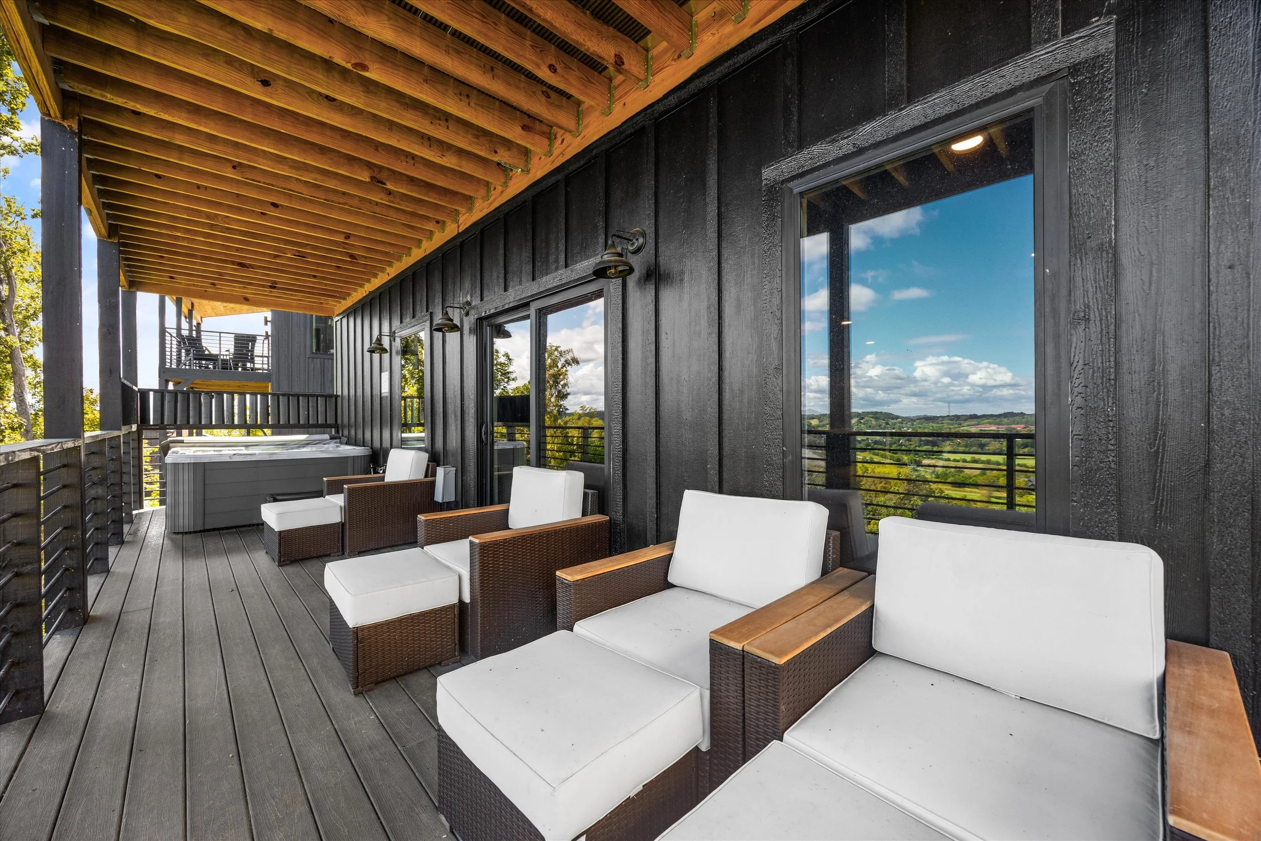 Outdoor balcony or patio area with wicker and white cushioned seating, black wooden walls, and a wooden ceiling, overlooking a scenic landscape with blue sky and clouds.