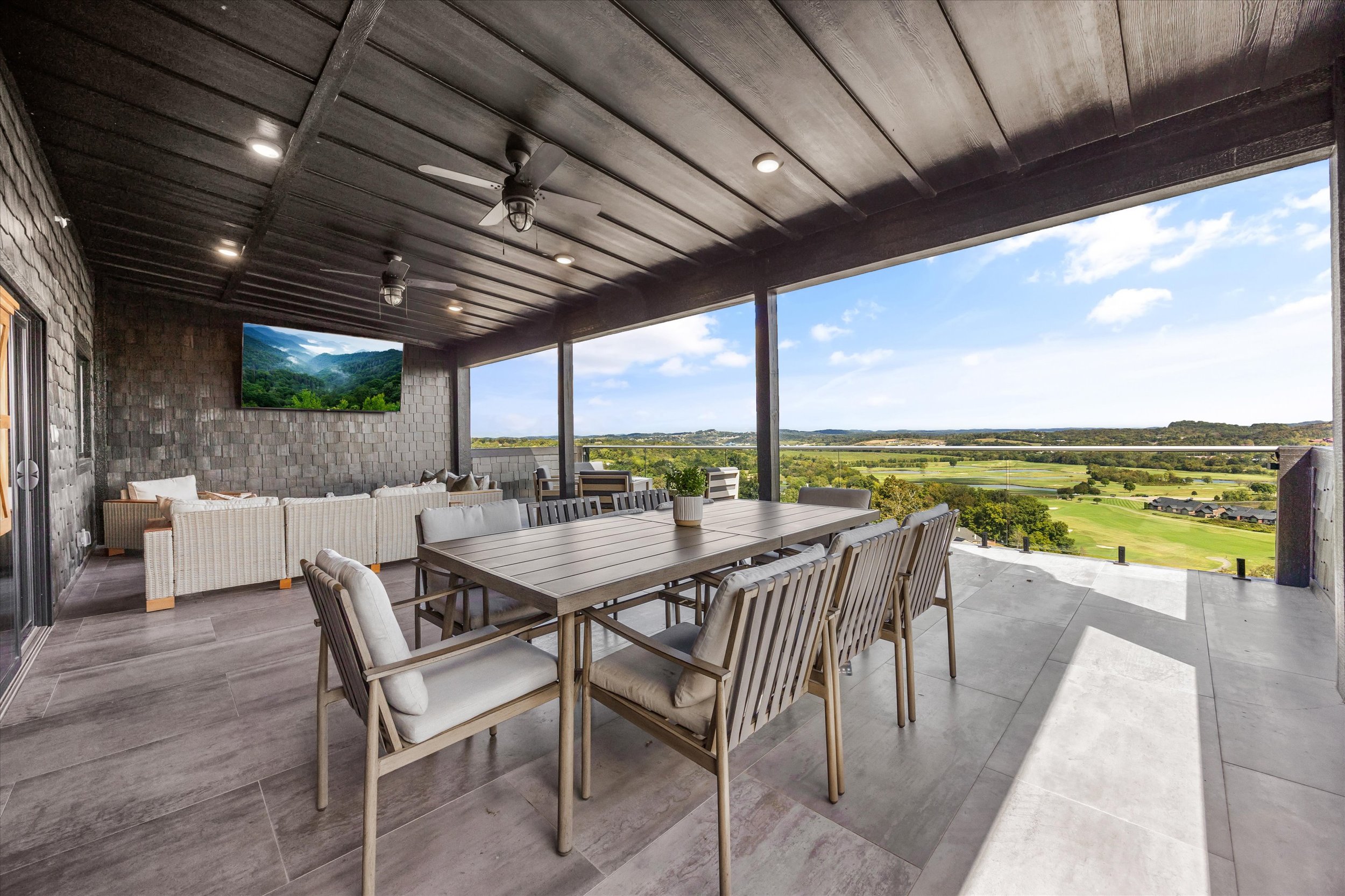 A spacious outdoor patio area with a large dining table and chairs, wicker lounge furniture, ceiling fans, and a scenic view of green fields and hills under a partly cloudy sky.