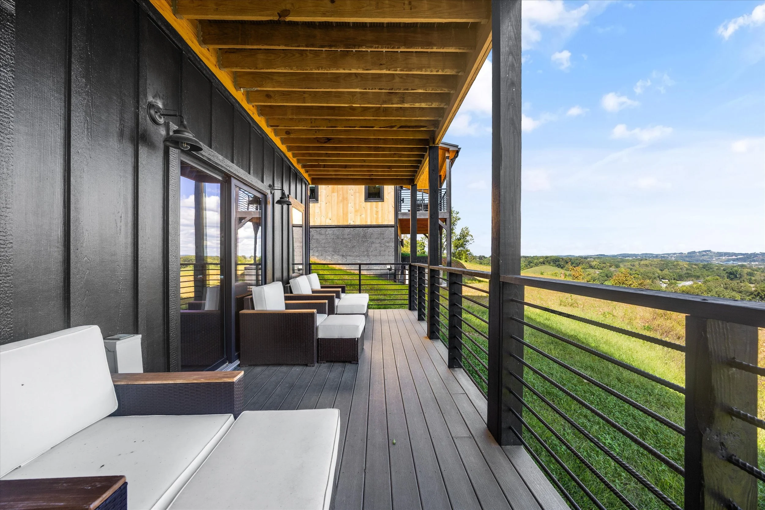 Balcony with black and natural wood finishes, outdoor seating with white cushions, overlooking green landscape under a bright blue sky with some clouds.