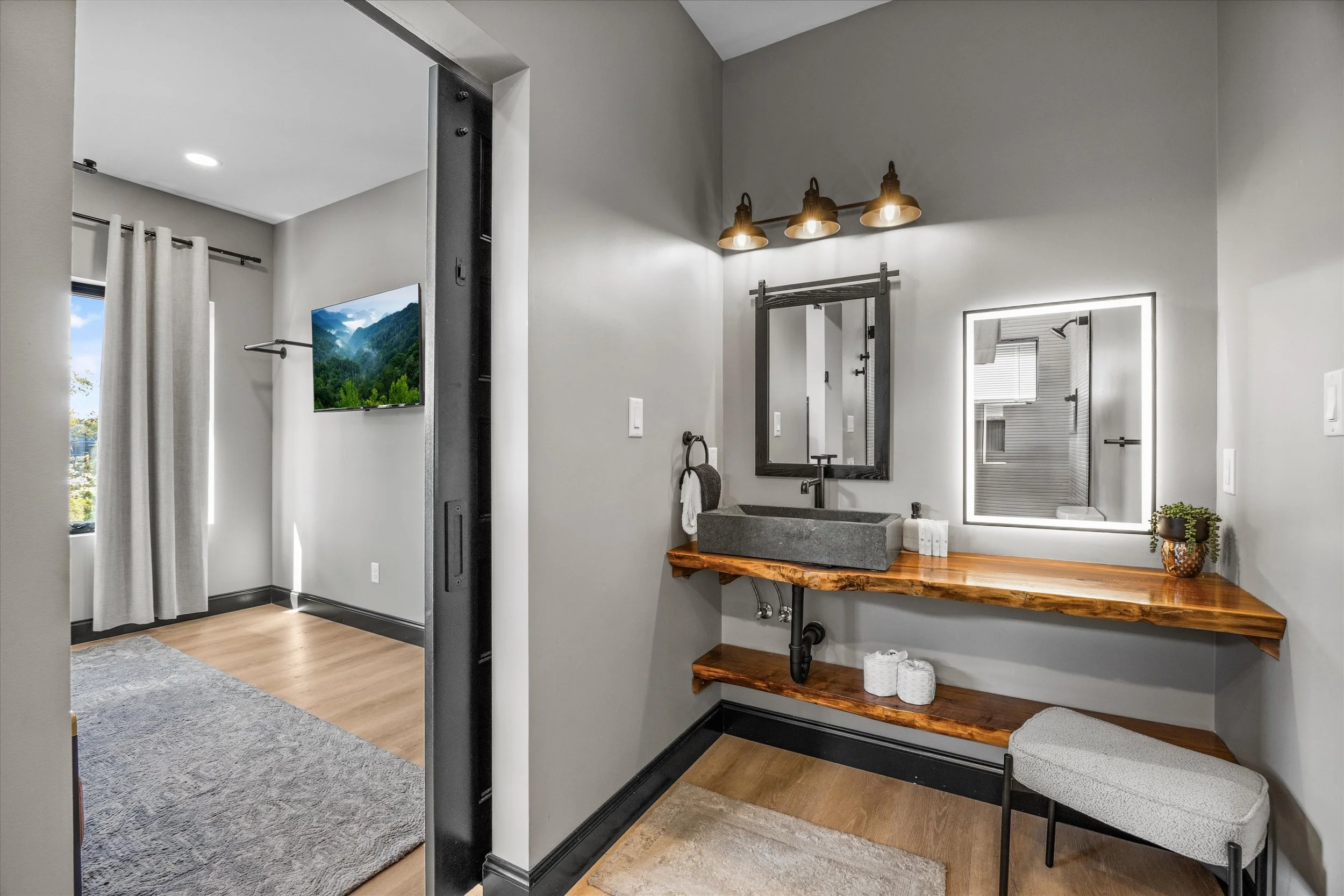 Modern bathroom with gray walls, a rectangular vessel sink on a wooden countertop, large illuminated mirror, and a black hand towel holder. Decor includes a potted plant and rolled towels. Visible adjacent room with window and television.