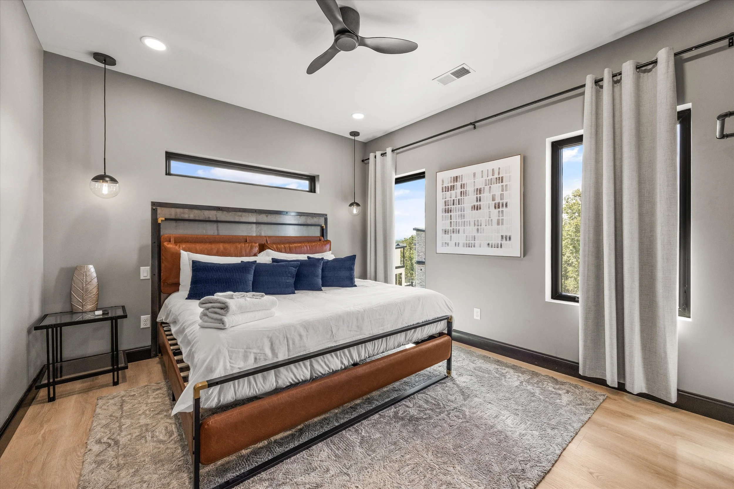 Modern bedroom with a bed featuring brown leather headboard and multiple navy blue pillows, two pendant lights hanging from the ceiling, gray walls, and large windows with white curtains, with a decorative vase on a black side table.