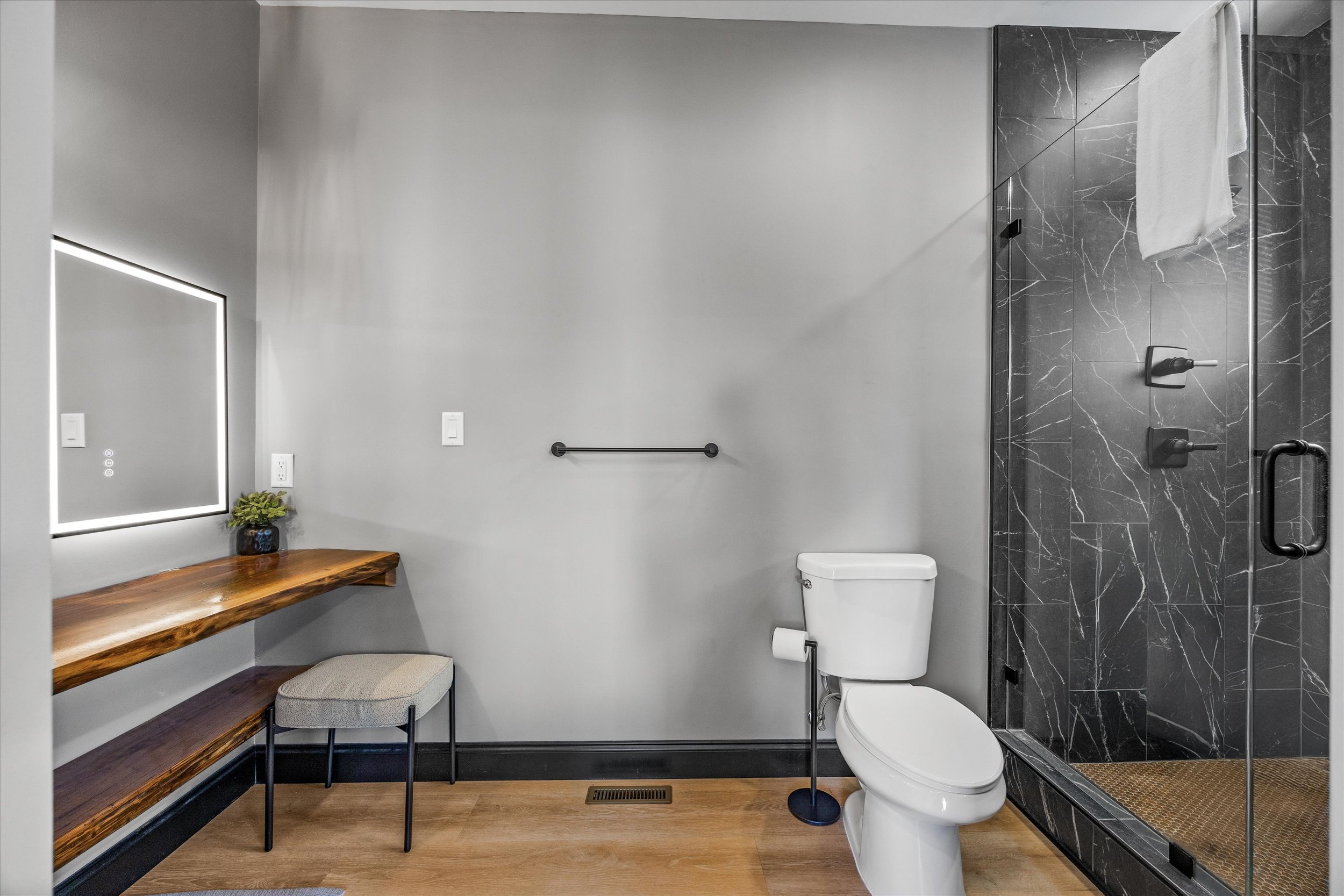 Modern bathroom with light gray walls and wood flooring. Features include a black shower with marble tiles, a white toilet, a black towel bar, a wooden bench with a stool, a small plant, and a rectangular mirror with LED lighting.