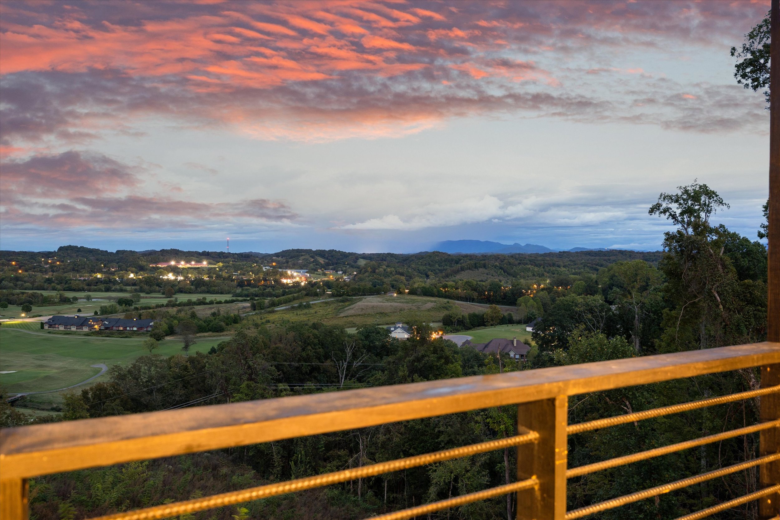 A scenic view of a sunset over a hilly landscape with scattered houses and trees, taken from a bridge with a yellow railing in the foreground.
