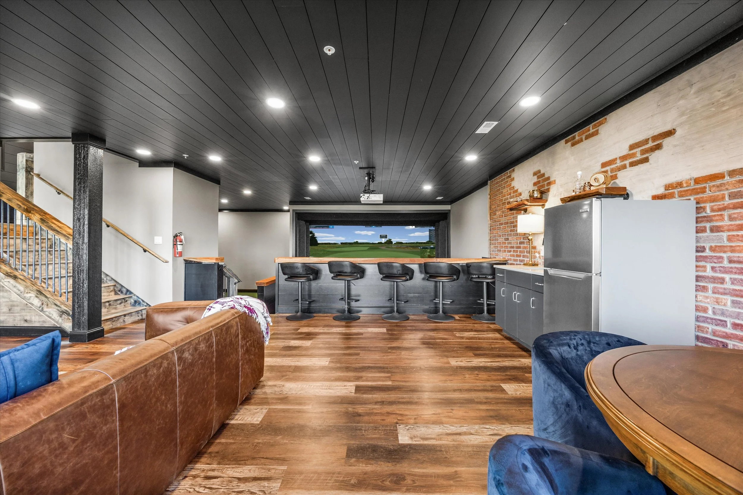 Modern living room with dark wooden ceiling, brick accent wall, leather sofa, and a bar area with stools facing a large window showing a green landscape.