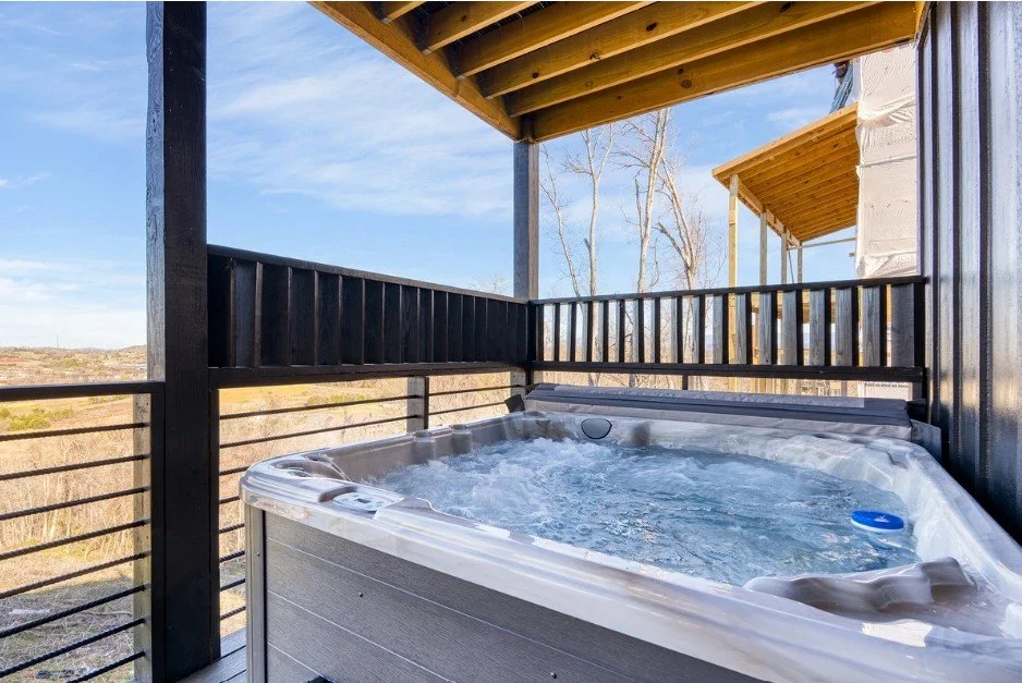 Hot tub with bubbling water on a balcony with black wooden railing and a wooden roof, overlooking a rural landscape with trees and open fields.