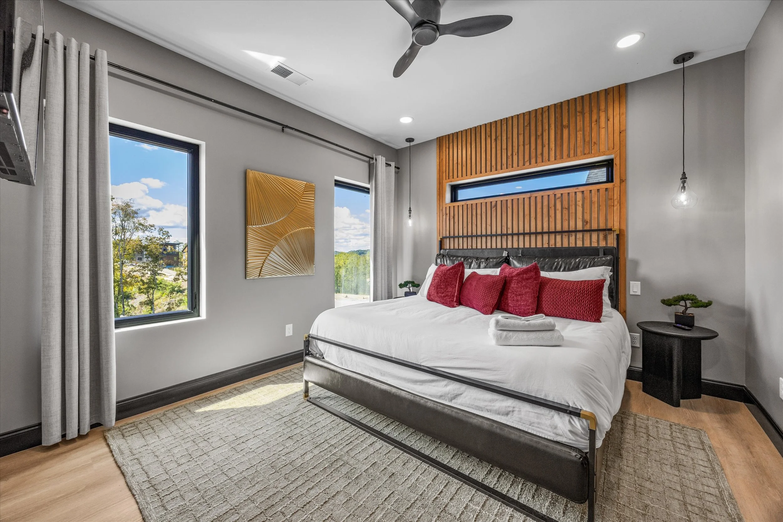 Modern bedroom featuring a bed with red pillows, a wooden accent wall behind the bed, and large windows with light gray curtains showing a view of trees and blue sky outside. There is a ceiling fan, two pendant lights, and a colorful wall art piece.