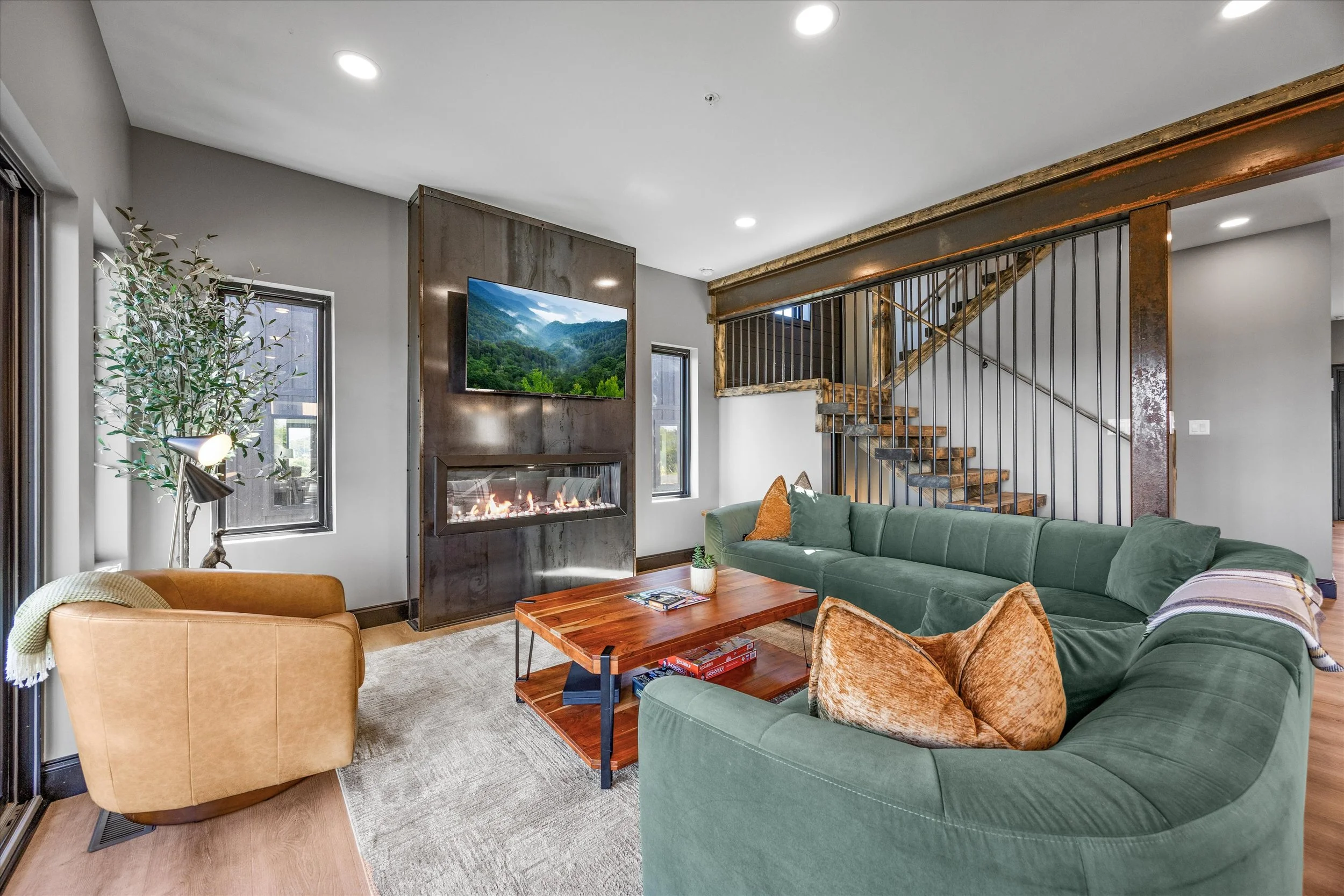 Living room with a green sofa, orange armchair, and wooden coffee table. Fireplace, wall-mounted TV showing mountains and greenery, window, and staircase with wooden steps and black metal railing.