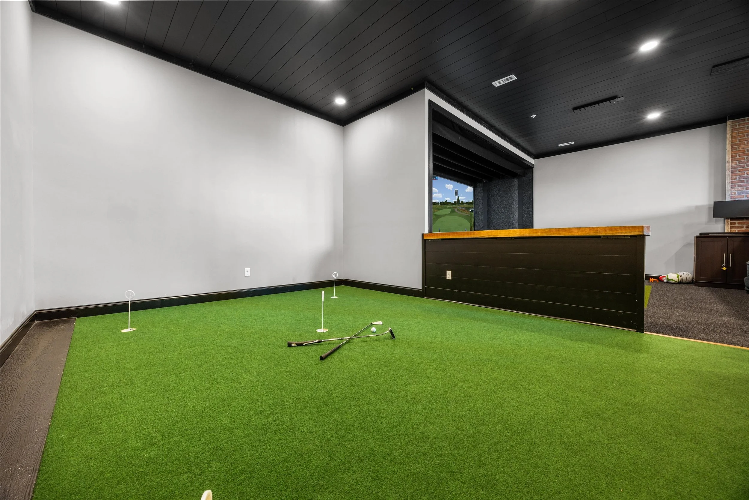 Indoor golf practice area with artificial green turf, golf balls, and golf clubs, featuring a window showing a golf course outside.