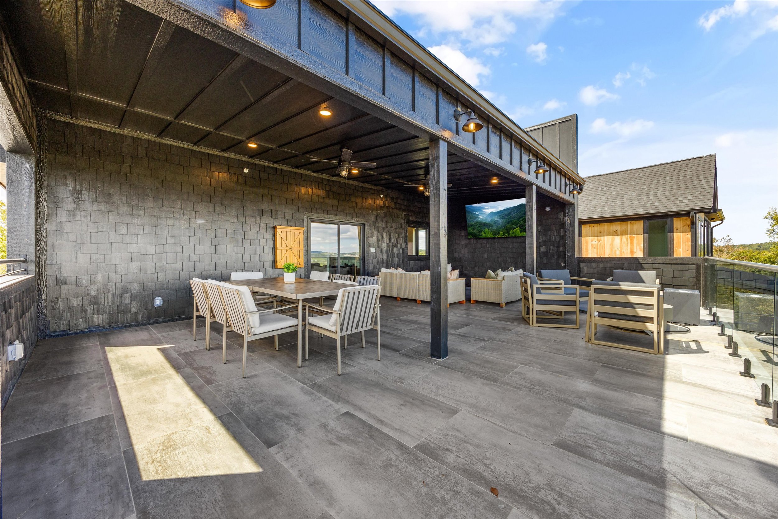 Outdoor balcony space with a dining table and chairs, seating area with sofas, and a view of mountains and trees in the background.