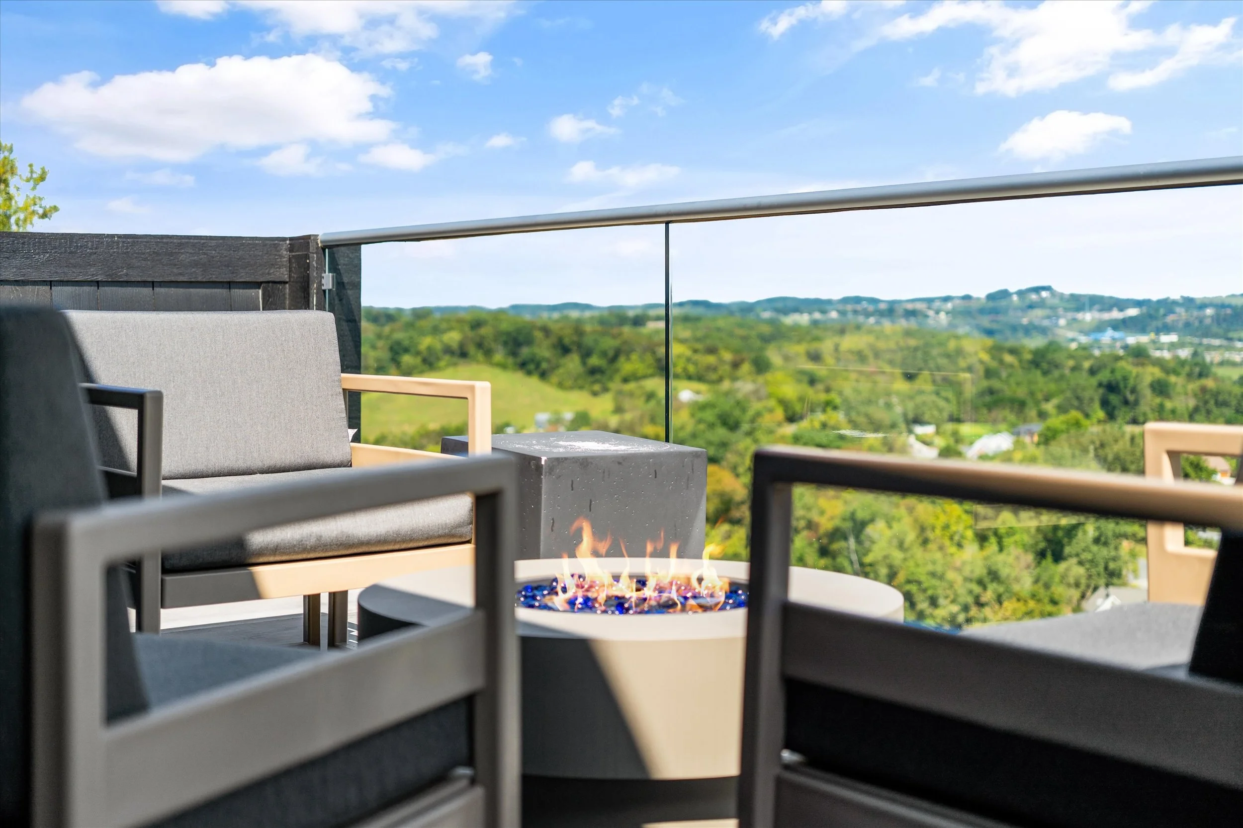 Outdoor balcony with chairs and a fire pit, overlooking a green hilly landscape under a partly cloudy sky.